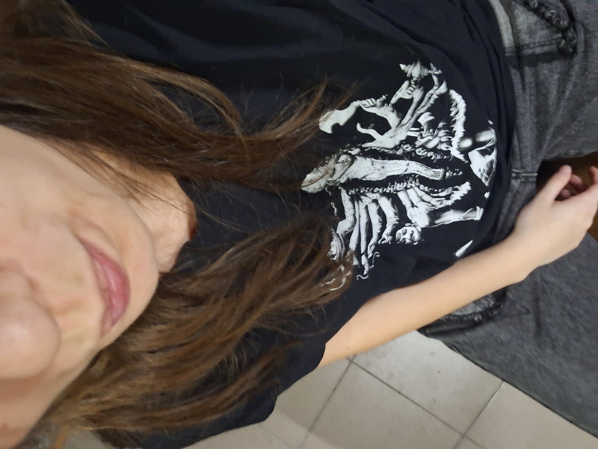 A selfie, primarily capturing the left side of a person's face, brown hair, and upper torso. They are wearing a black t-shirt with a white graphic print featuring skeletal figures. The person is also wearing gray jeans. The background shows a light-colored tiled floor. No information indicates that the image was taken in Panagyurishte, Bulgaria.