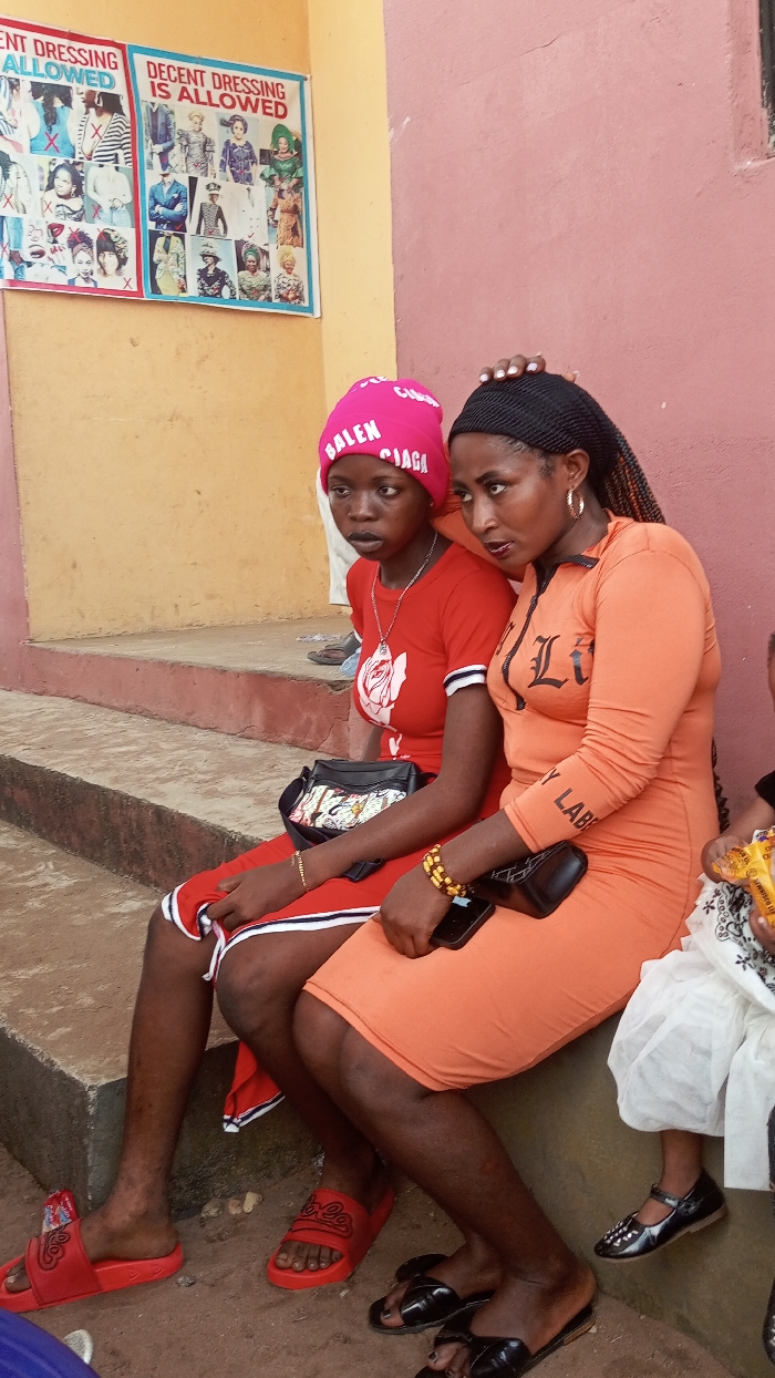 Two women sit close together on concrete steps. One wears a pink beanie, red t-shirt, red shorts, and red slides. The other wears an orange dress with a zipper, gold hoop earrings, and black sandals. The woman in orange places her hand on the other's head. A "Decent Dressing is Allowed" poster hangs above on a yellow wall, and a pink wall is to the right. Part of a child wearing black shoes and a white dress is visible on the right edge.