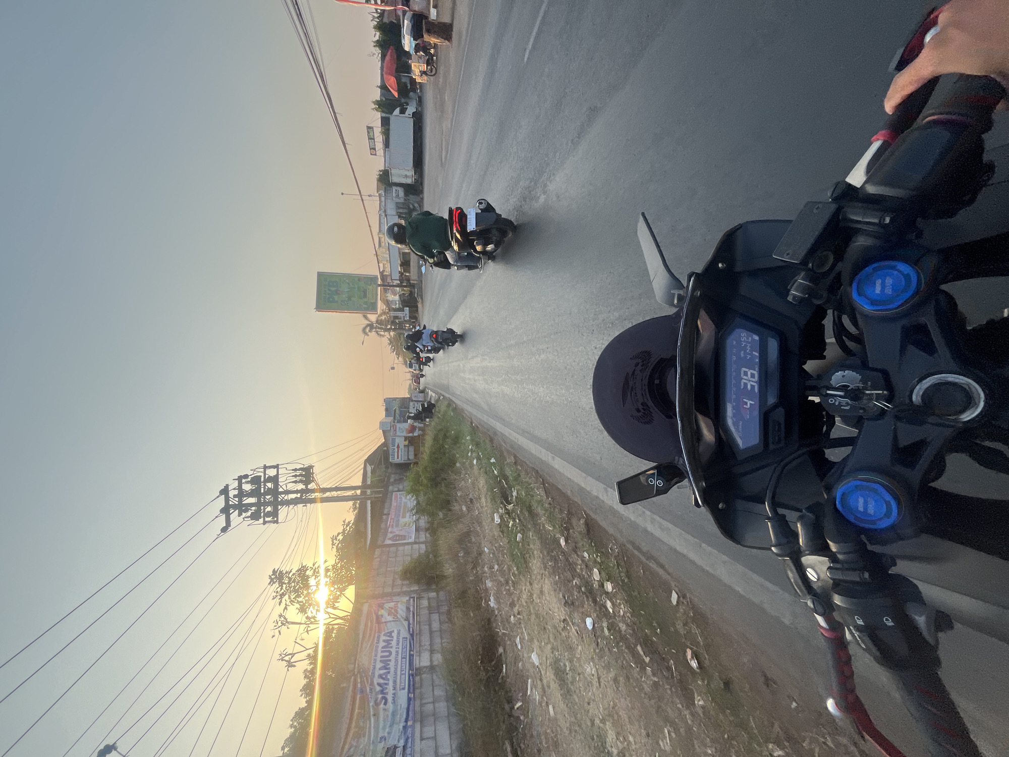The image is a first-person perspective shot from a motorcycle in motion on a paved road, likely in the vicinity of Sengonbugel, Indonesia, given the provided context. The time of day appears to be late afternoon or early evening, with the sun setting low in the sky, casting a warm, golden light. The road is lined with vegetation and some scattered trash on the left side, while the right side features buildings, signs, and other vehicles.
In the foreground, the handlebars and dashboard of the motorcycle are visible, with a digital display showing "4 38" and other unreadable symbols. The rider's hands are on the handlebars, indicating they are actively riding. The road itself has visible lane markings and is reflecting the light from the setting sun, suggesting it might be damp.
Further down the road, several other motorcycles are visible, some with riders. One motorcycle in the middle ground has a rider wearing a green jacket. In the background, there are buildings, electrical poles with numerous wires, and what appear to be trucks and other vehicles. A large billboard with text in Indonesian is visible on the left side, and a smaller green billboard is further down the road on the right. The overall environment suggests a semi-urban or peri-urban setting during a motorcycle ride.