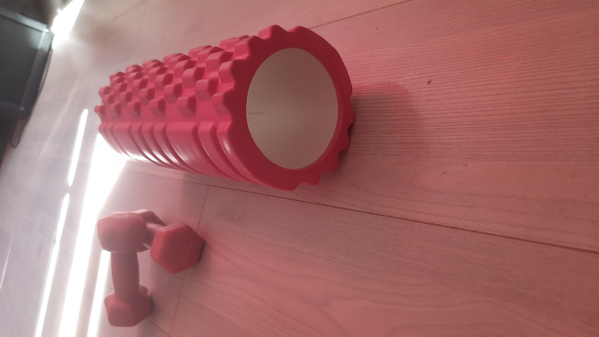 A pink foam roller and two pink dumbbells are arranged on a light pink wooden floor. The foam roller is in the foreground, positioned vertically and to the right of the frame. It has a textured surface with raised bumps. The dumbbells are in the background, to the left of the frame, one slightly behind the other. The scene is lit by bright, horizontal light sources from the top left, casting soft shadows on the floor. The environment appears to be indoors. There is no indication of the location being in Desnogorsk, Russia.