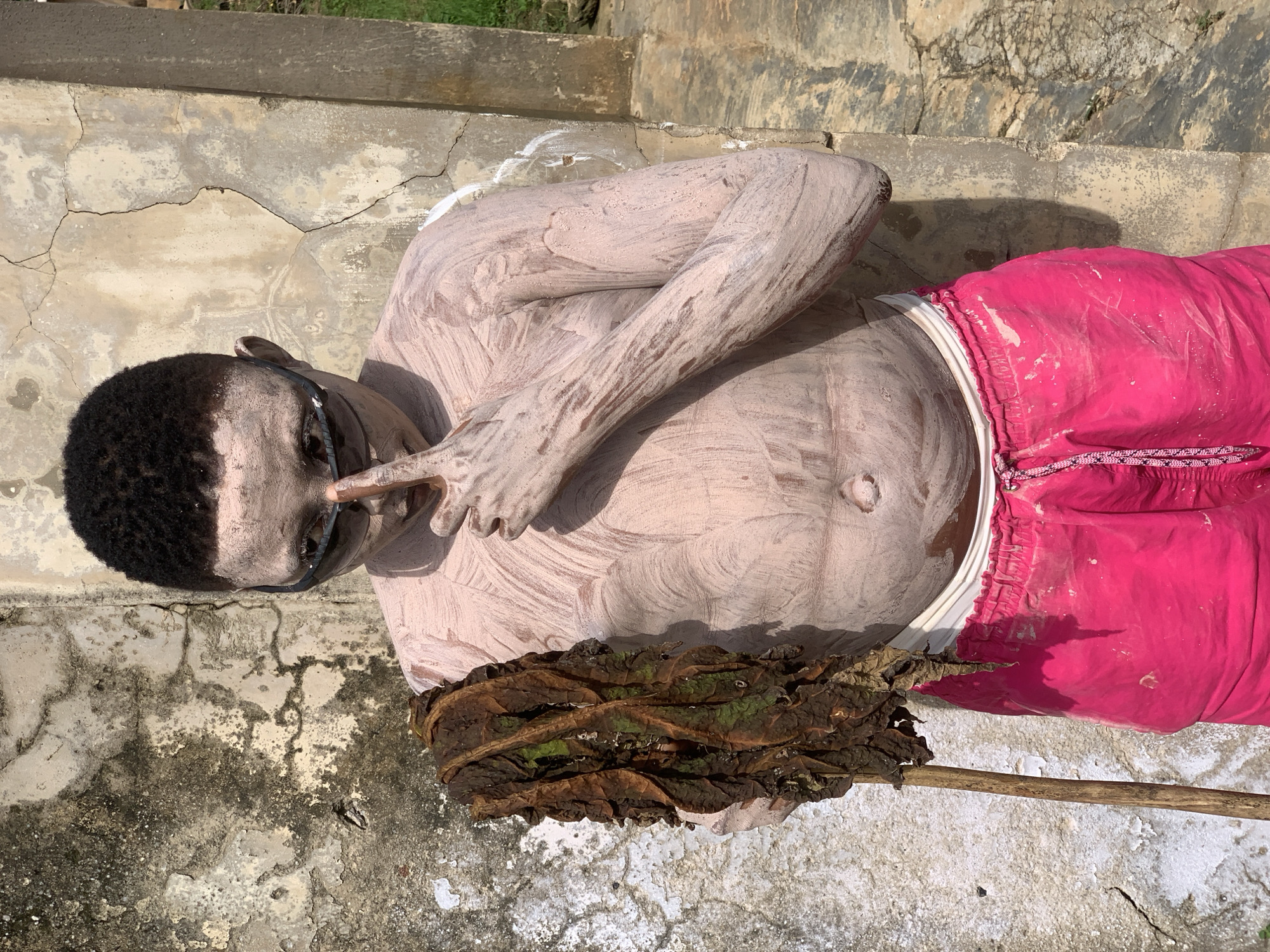 A person lies on concrete steps, covered in a light pinkish-tan paste or paint. They are wearing bright pink shorts. Their hair is dark and short, and they have on dark glasses. One arm is bent, with fingers touching their face near their nose. Next to them, on the steps, is a bundle of dry, brown and green leaves attached to a stick, resembling a broom. The concrete steps are worn and cracked, with patches of dirt and some white residue. In the background, a wall with similar concrete texture and some greenery are visible. The lighting suggests it is daytime, with shadows cast on the steps. The overall scene appears to be outdoors in an urban or semi-urban setting in Ikorodu, Nigeria. No visible text is present in the image.