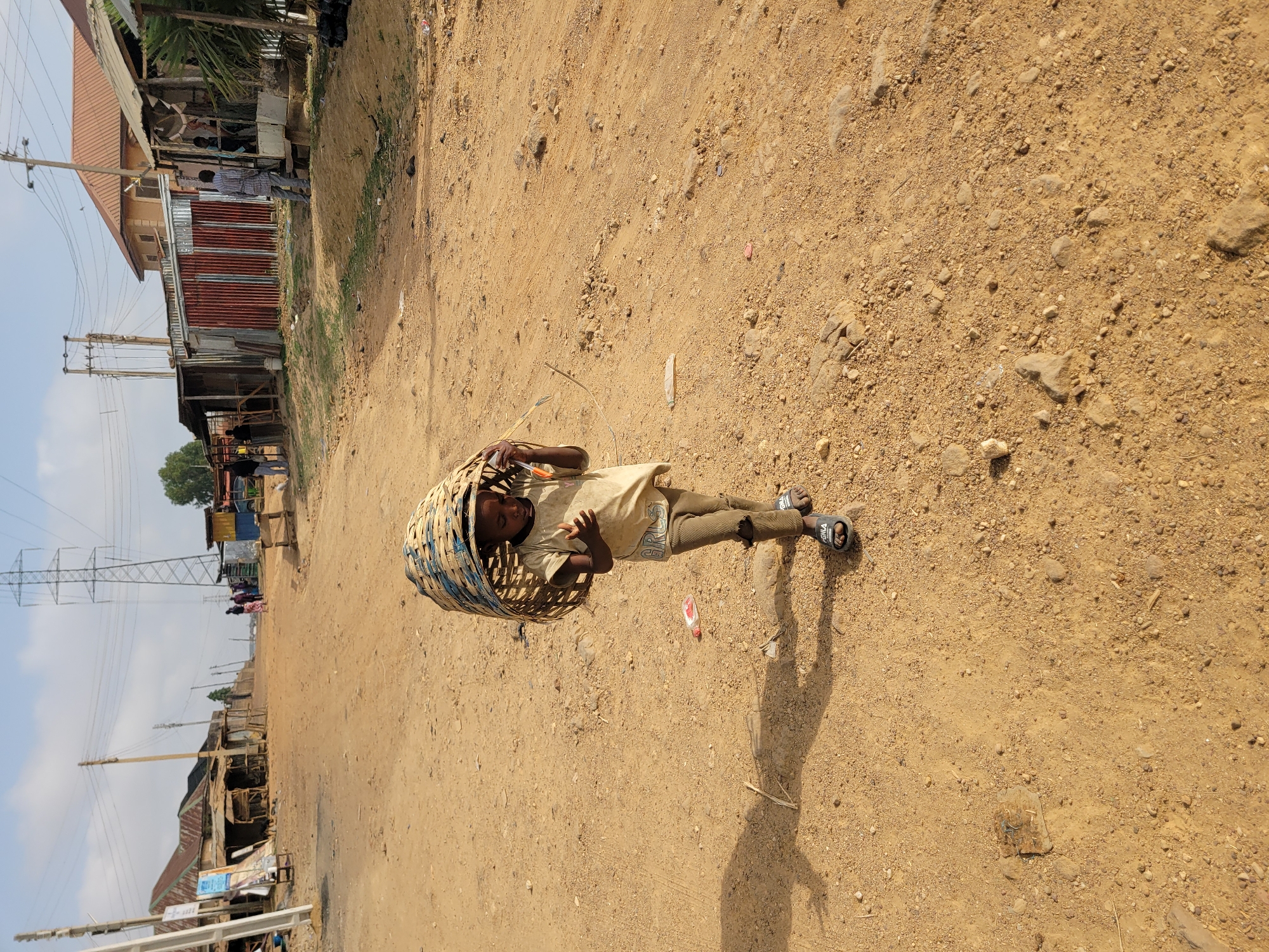 A young person carries a basket on their head in Jos, Nigeria. The dirt road has small rocks. Buildings and power lines line the road in the background. The young person is wearing a light-colored shirt, pants, and sandals.