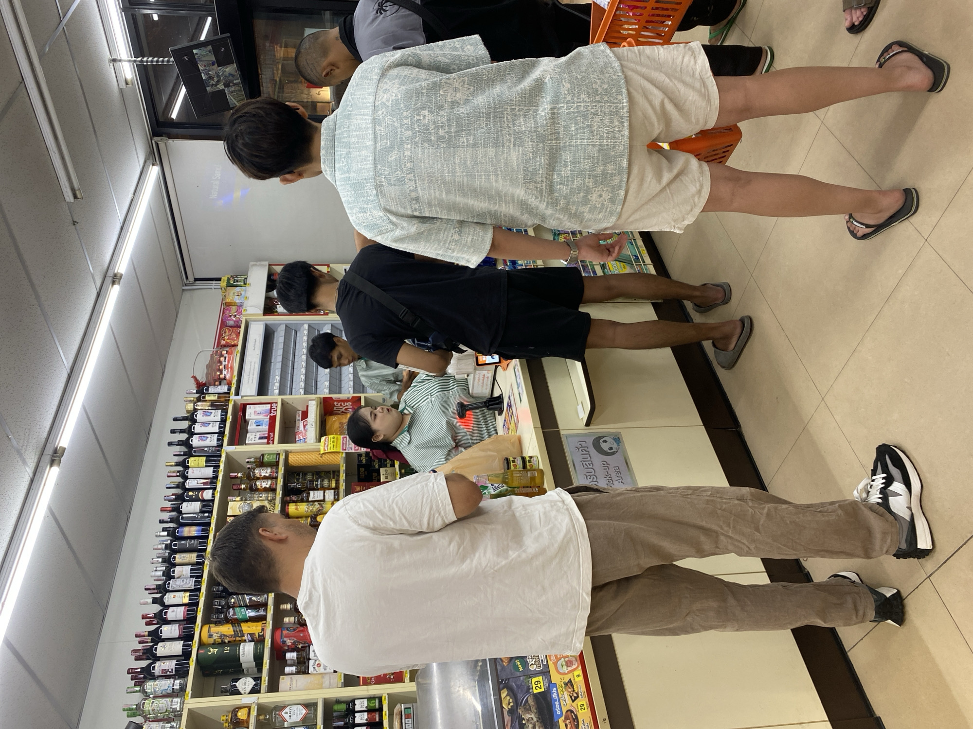 This media file shows the interior of a convenience store in Baan Bo Phut, Thailand. Several people are visible, including customers and a cashier.
In the foreground, a person wearing a white t-shirt and brown pants is standing, facing away from the camera. To their left, another person in a light blue patterned shirt and white shorts is also visible. Behind them, a person in a black t-shirt and shorts is standing. Further back, a cashier wearing a green and white striped shirt is attending to a register. The shelves in the background are stocked with various items, including bottles of alcohol and other products.
The store appears to be well-lit, suggesting it is open for business. There is visible text in both English and Thai on signs and product labels. Some of the text includes "Natural Samui," "Pick-up Area," and product names like "Jim Beam" and "Blend." The overall scene depicts a typical shopping environment in a convenience store. The time of day and weather are not discernible from the image. The people's expressions are not clearly visible, but the general atmosphere appears to be that of a transaction or shopping.