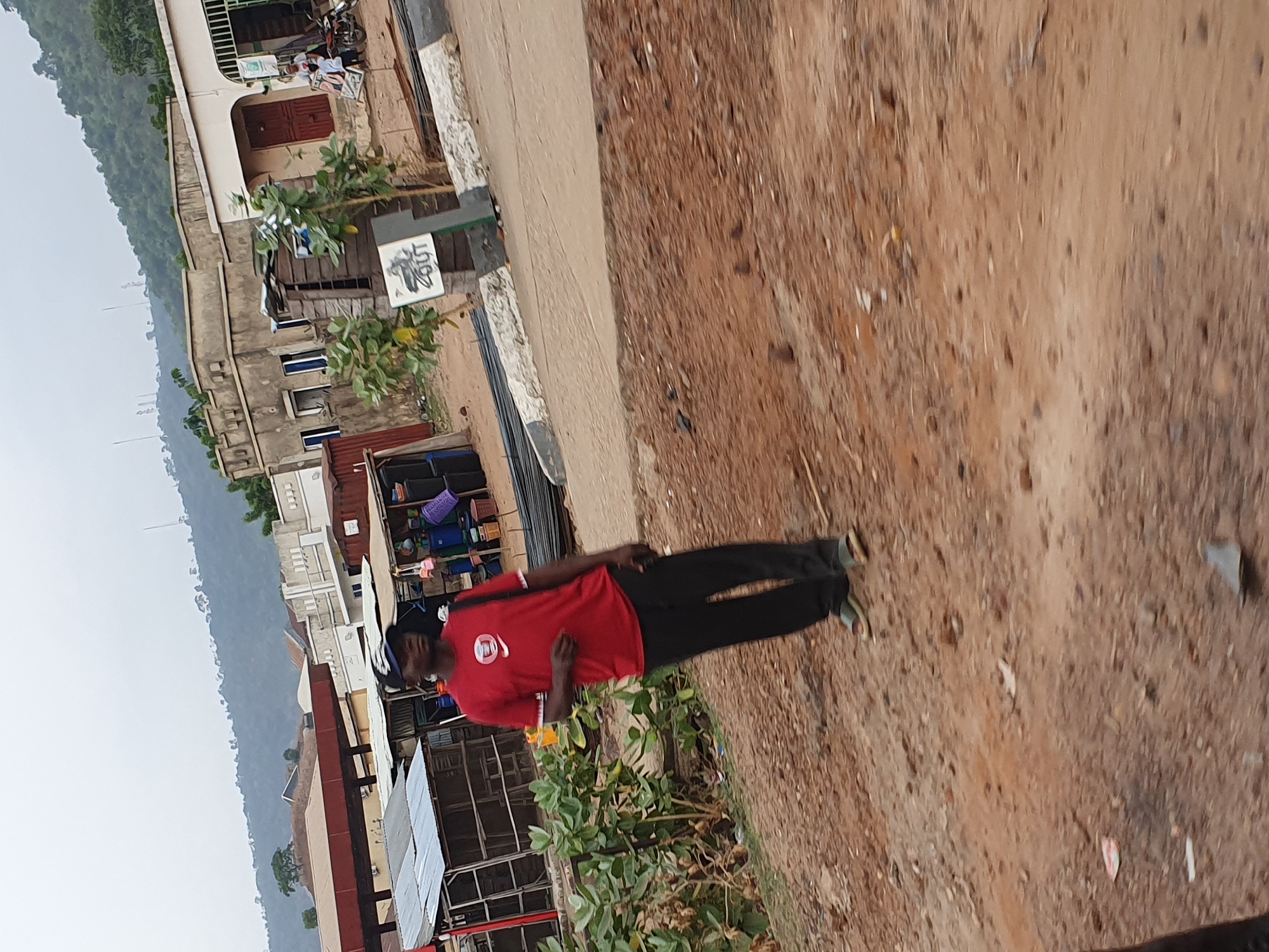 This full-shot image captures a man standing on a reddish-brown dirt path or road, flanked by buildings and natural landscape under a hazy sky. The image is presented rotated 90 degrees clockwise.
The main subject is a man standing slightly left of center. He is dark-skinned with short dark hair, wearing a red short-sleeved t-shirt with a white logo resembling the Nike swoosh and additional text (unreadable) on the front. He is also wearing black trousers and light-colored sandals. His posture is relaxed, and he appears to be looking towards the left side of the frame with a neutral expression.
The immediate foreground and midground feature a rough dirt path or road, scattered with small stones and sparse, dry vegetation. To the man's left, a small green bush is visible. Behind him, a series of simple, multi-story concrete buildings line what appears to be a street. These buildings have various architectural details, including visible windows and doors, some with a slightly worn appearance. One building features a distinct red door, and another has various items stacked outside, possibly indicating a market stall or shop, including colorful plastic basins. A white sign with black stylized text reading "50hz" is clearly visible near one of the buildings. A motorcycle is parked further back near another structure.
In the distant background, a range of green, tree-covered hills or mountains rises, extending across the horizon. The sky above is overcast or hazy, suggesting a soft, diffused daylight, possibly early morning or late afternoon. The overall scene portrays a typical street environment in a semi-urban or rural setting, consistent with the given location of Lokoja, Nigeria.