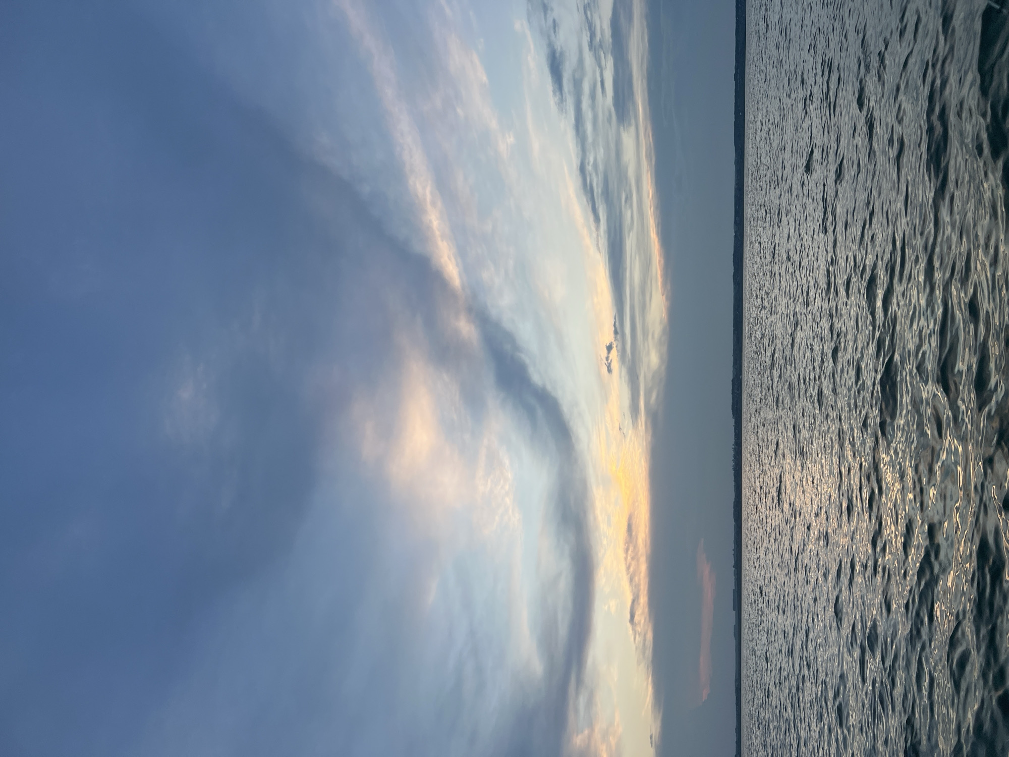 This is a vertical, eye-level, medium shot photograph of a body of water under a cloudy sky at dusk. The water occupies the lower half of the frame, with small ripples reflecting the faint light from the sky. The horizon line is visible as a dark, indistinct band of land, possibly the city of Novopichugovo, Russia, separating the water from the sky. The sky is filled with a variety of clouds, ranging from dark, heavy masses to wispy, lighter formations. Patches of pale orange and yellow light are visible through breaks in the clouds, suggesting the setting sun. The overall atmosphere is serene and calm. There are no people or specific objects visible in the image.
