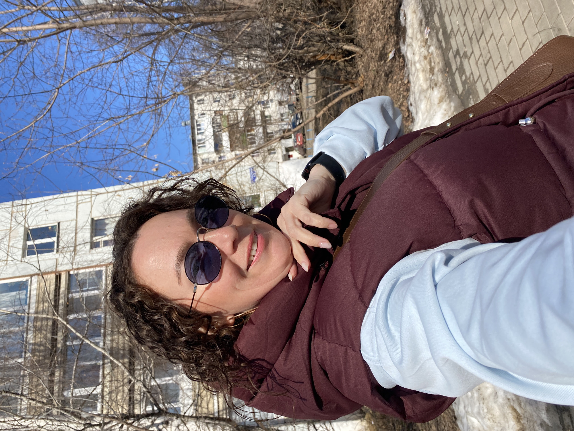 The image is a selfie of a woman wearing sunglasses and a burgundy puffer vest over a light blue long-sleeved shirt. She has curly brown hair and is smiling. The background includes bare trees and a light-colored building, which appears to be a multi-story residential structure. Some snow is visible along the ground and a brick sidewalk. Based on the visual cues and context, the location could be Chaikovsky, Russia.