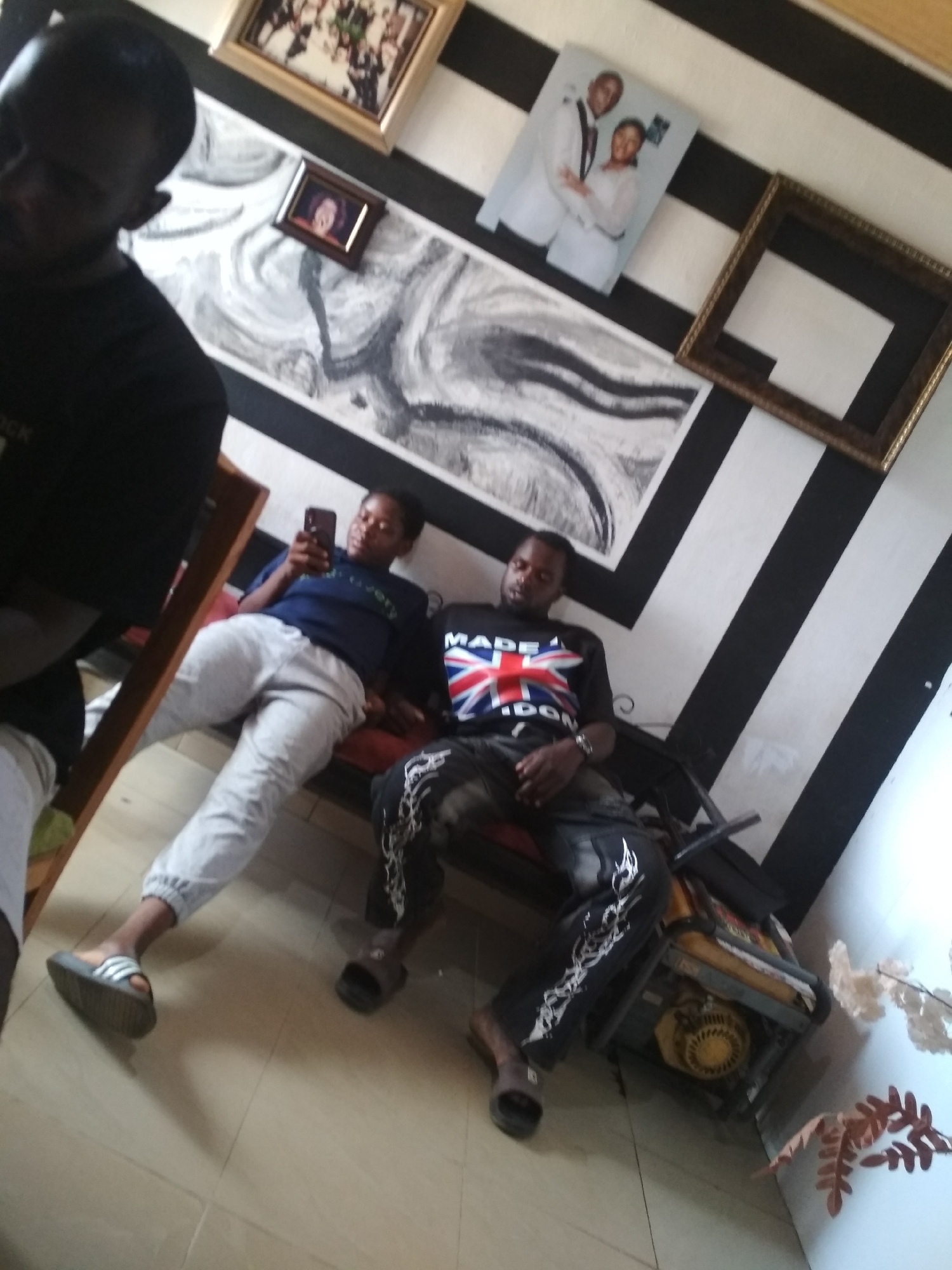 A slightly high-angle medium shot depicts three people indoors, likely in Unknown, Unknown. The back wall has alternating black and white diagonal stripes. Five framed pictures hang on the wall, including one with a couple in formal attire. Two of the people are seated. The seated woman is looking at a cell phone. The seated man wears a "Made In London" t-shirt with the Union Jack flag. He is wearing dark pants with white designs down the side. There is an orange generator next to him. A third person is partially visible on the left. All three are Black.
