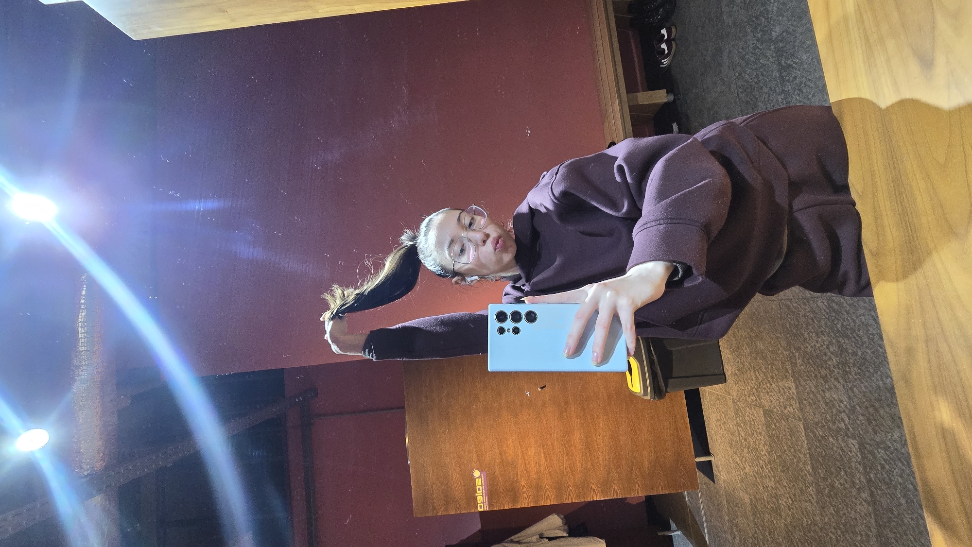 A young woman is taking a selfie in a mirror. She is wearing glasses, a purple hoodie, and has her hair in a high ponytail. She is holding a light blue smartphone, likely taking the photo with it. The background behind her features a red wall and a wooden desk. The mirror reflection also shows a window with bright light shining through it. The location appears to be indoors, and the surrounding environment doesn't clearly indicate the city of Stara Zagora, Bulgaria.