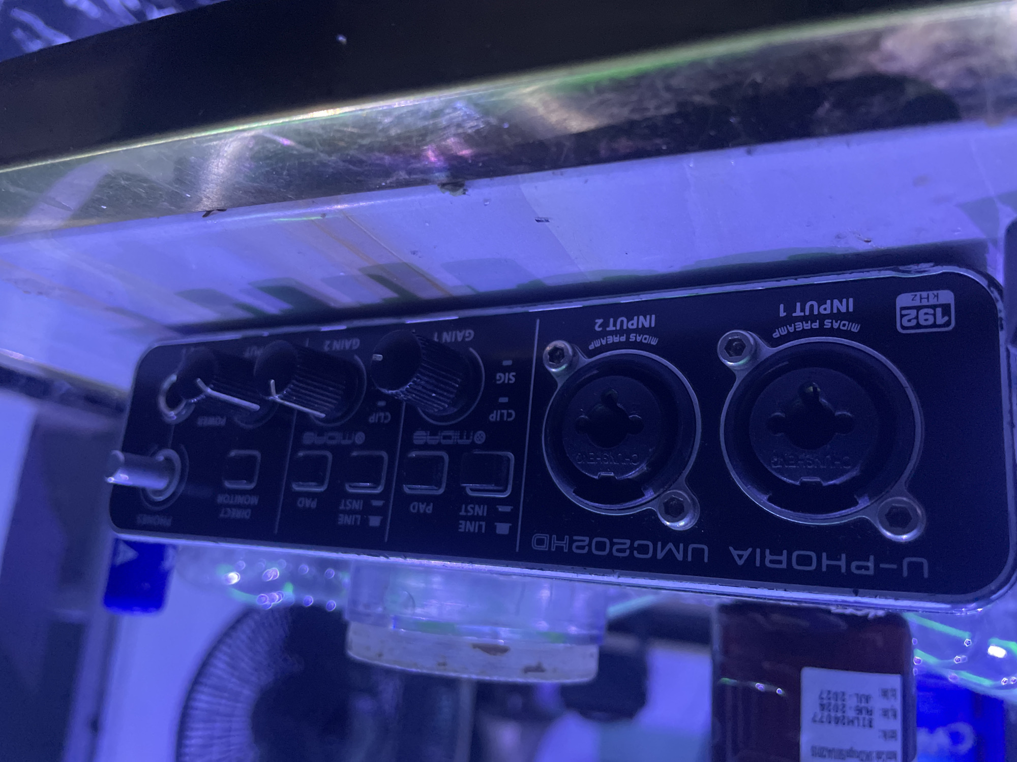 The media file displays the front panel of a black Behringer U-PHORIA UMC202HD audio interface. The device is prominently featured, illuminated by strong blue artificial light that creates reflections on its surfaces and the surrounding environment.
The main subjects are the various controls and inputs of the audio interface. On the right, there are two combo XLR/TRS inputs labeled "INPUT 1" and "INPUT 2," both featuring "MIDAS PREAMP" technology. To the left of these inputs are two large rotary "GAIN" knobs (GAIN 1, GAIN 2) for adjusting input levels, each accompanied by "SIG" (signal) and "CLIP" indicators. Below the gain controls are toggle switches for each input, allowing selection between "LINE" and "INST" (instrument) inputs, and a "PAD" switch for attenuating the signal.
Further to the left, there is a "DIRECT MONITOR" button, a "POWER" indicator, and a "PHONES" (headphones) output with its own volume knob. The top right corner displays "192 KHZ," indicating the maximum sample rate. The brand name "U-PHORIA UMC202HD" is visible along the bottom edge of the device. Connectors inside the input jacks bear the text "CHUNSHENG."
The setting appears to be within or behind a clear, reflective surface, possibly an acrylic or glass enclosure, given the blue light and reflections that suggest an aquarium or similar setup. There are faint bubbles or debris on the clear surface above the device. A partially visible, clear plastic stand supports the audio interface. In the lower right, a blurred label is visible with text including "31LM24077" and "AJL-2027." The background shows blurred objects, possibly a fan or other electronic components, consistent with an indoor, technical setup. No specific activity is occurring; the device is static.