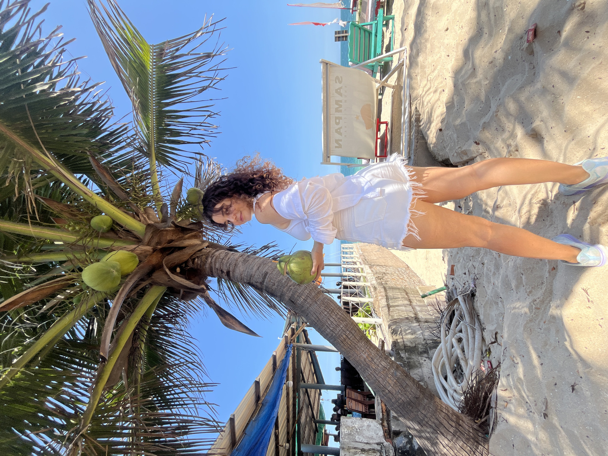 The image depicts a woman standing on a sandy beach in Phan Thiet, Vietnam. She is wearing a white off-the-shoulder top and white denim shorts, holding a green coconut in her right hand. The woman is positioned in front of a leaning palm tree, which has several coconuts growing on it. In the background, there is a beachside establishment with a blue tarp roof, tables, and chairs, suggesting a restaurant or bar. Further in the background, beach chairs and umbrellas are visible, along with the ocean. The sand is light brown, and the sky is clear and blue. There are also some coiled white hoses and debris on the sand near the base of the palm tree.