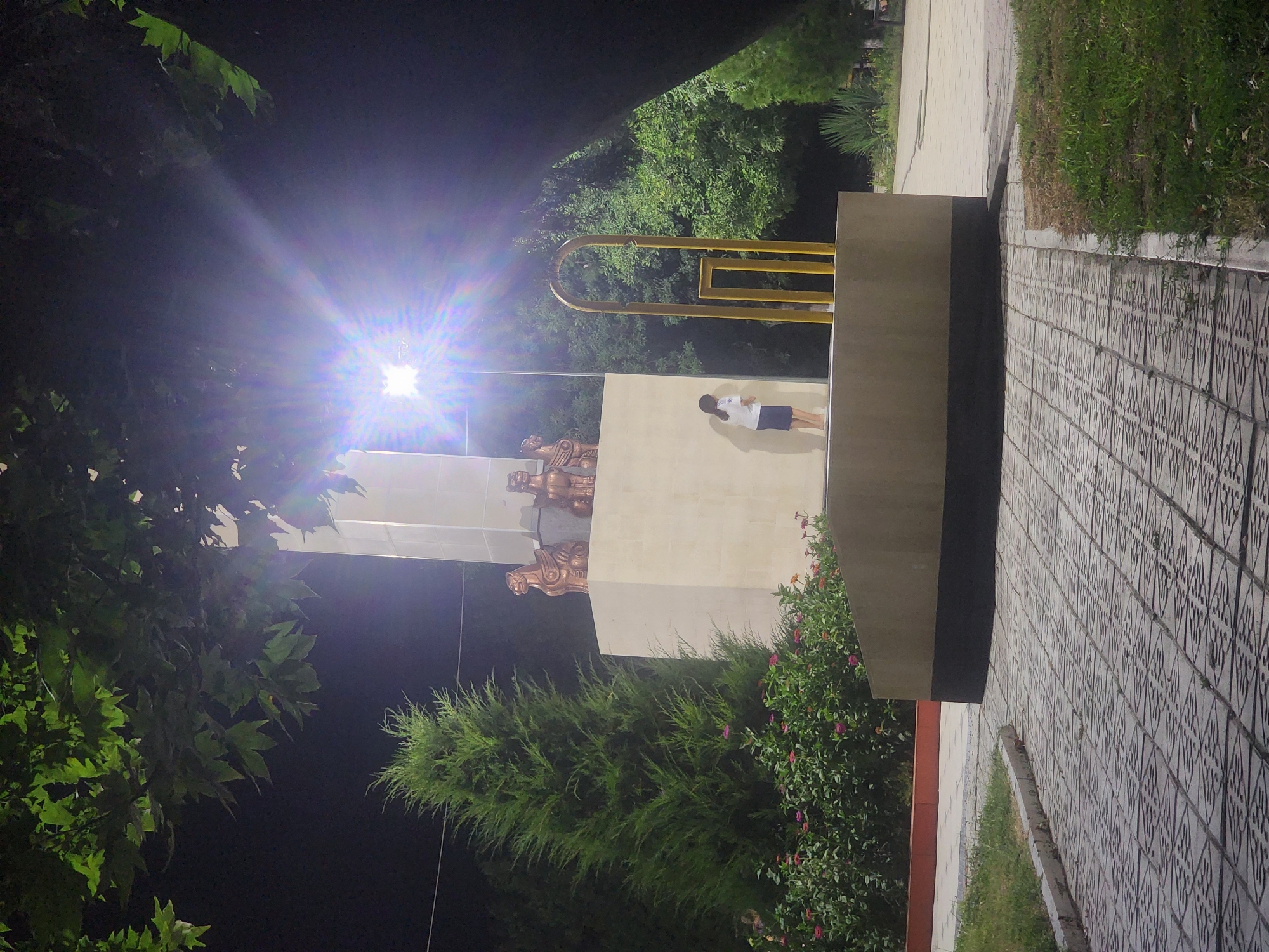 A young girl with dark hair stands with her back to the camera, wearing a white t-shirt and a blue skirt. She is positioned on a raised platform in front of a tall, light-colored monument. The monument has bronze sculptures of mythological creatures at its top. To the left of the monument is a tall, cone-shaped evergreen tree, and in front of that is a bed of flowering plants with pink and orange blossoms. To the right of the girl and the monument is a yellow, abstract sculpture. The scene is illuminated by a bright light source positioned above and to the right of the monument, causing lens flare. The ground in front of the monument is paved with interlocking gray tiles. In the background, there are more trees and foliage. The overall setting appears to be a park or public square in Jetisay, Kazakhstan, likely in the evening or at night due to the dark sky and artificial lighting.