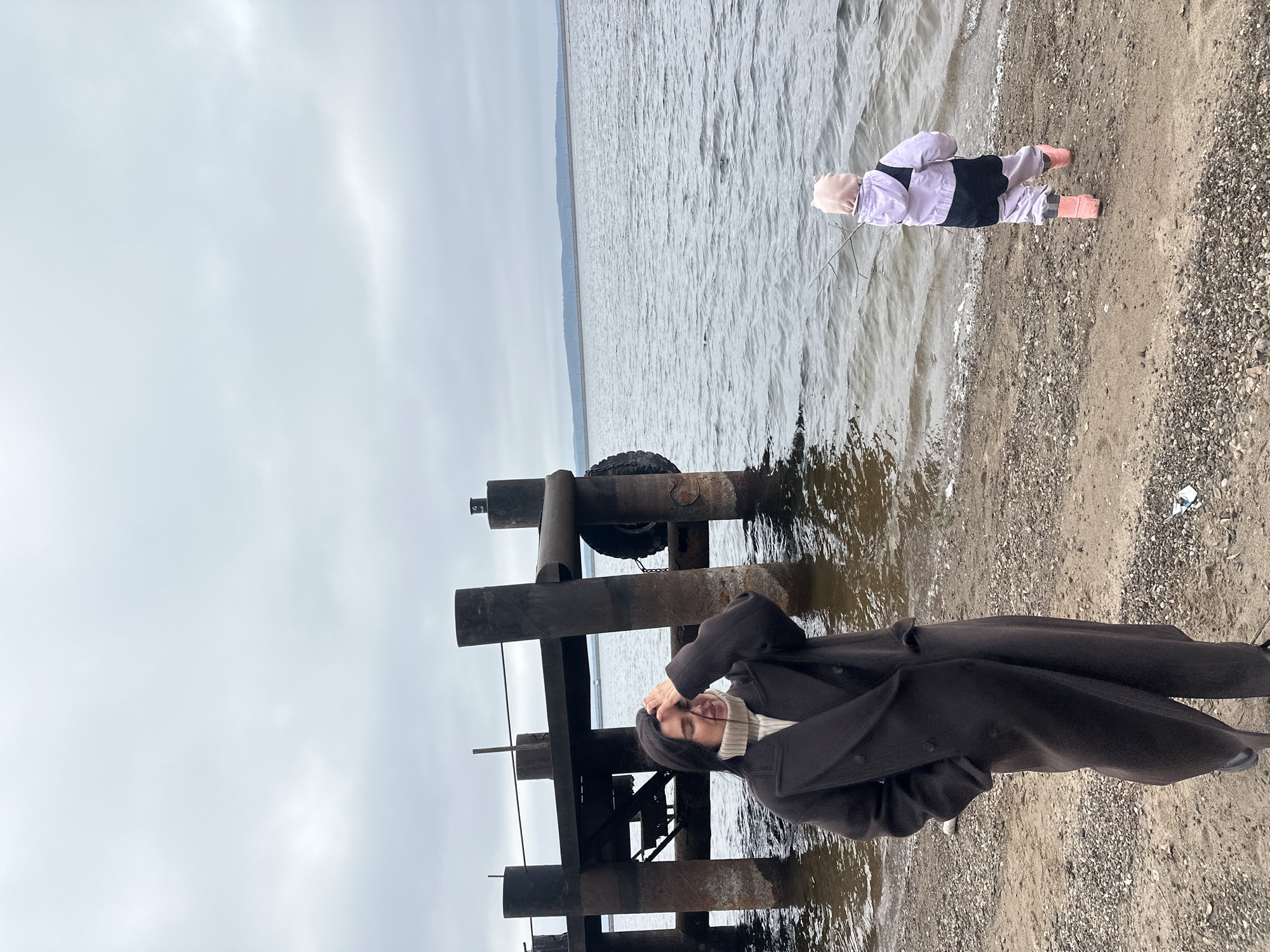 The image captures an outdoor scene by a large body of water under an overcast sky. In the foreground, positioned to the left of the frame, a person stands near a robust, weathered wooden pier. This individual appears to be an adult, dressed in a long, dark brown coat over a light-colored turtleneck sweater, with dark hair partially obscured by a raised hand near their head, possibly adjusting hair or a head covering.
Further to the right, a small child is visible at the very edge of the water, facing away from the camera. The child wears a light purple jacket, dark pants, and bright pink rain boots, with a light-colored hat. A small stick or branch lies on the sand near the child's feet.
The body of water, likely a lake or a wide river, stretches towards a faint horizon where distant land is barely visible through the haze. Its surface is ruffled with small waves. The immediate shoreline consists of a mix of sand and small pebbles or gravel. The wooden pier is constructed from thick, dark timber beams, and a black tire hangs from one of its supports, likely serving as a fender. The sky is a uniform, pale gray, indicating an overcast or cloudy day.
The attire of both individuals suggests cool or chilly weather. The scene conveys a calm and somewhat serene atmosphere. There is no visible text in the image. Given the location context, this scene likely depicts a spot along the Kama River or Votkinsk Reservoir in Chaikovsky, Russia.