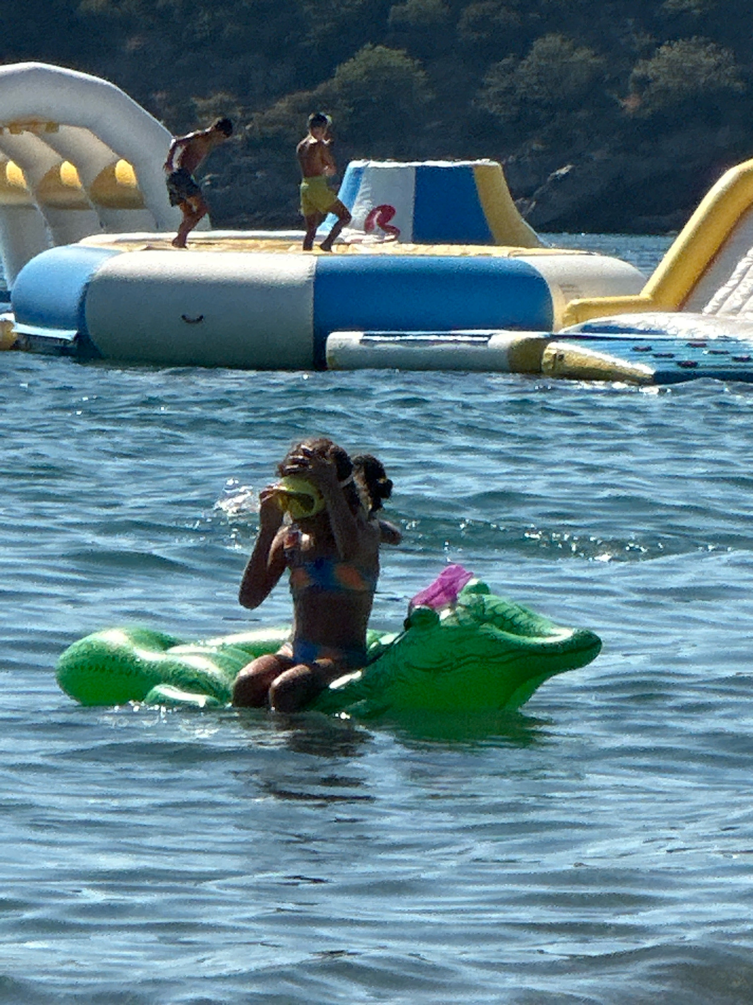 In the waters of Boreti, Montenegro, a girl wearing a colorful bikini is seated on a green inflatable alligator. She is holding a yellow object to her face, possibly a toy or a piece of fruit. In the background, a large inflatable water park structure with slides and climbing walls is partially visible. Two boys are on the water park; one is wearing dark swim trunks and the other is wearing yellow shorts. The water is choppy and reflects the sunlight. The background shows a tree-covered hillside.