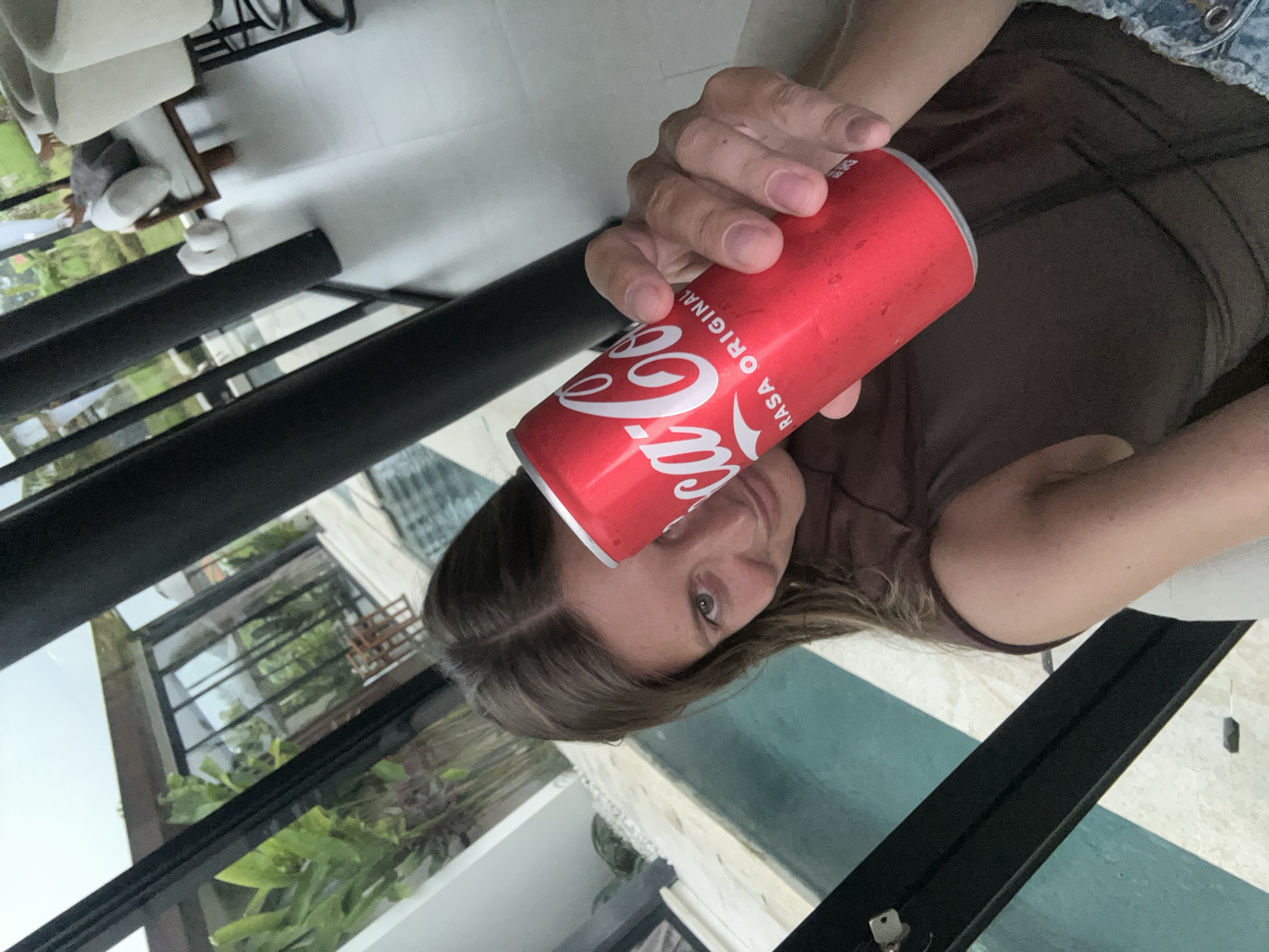 A woman is holding a red Coca-Cola can. The location appears to be in the Ubud District, Indonesia, based on the view through the windows, which reveals tropical foliage and architectural styles typical of the area. The woman is wearing a brown top. There are white chairs and a black metal structure visible in the background.