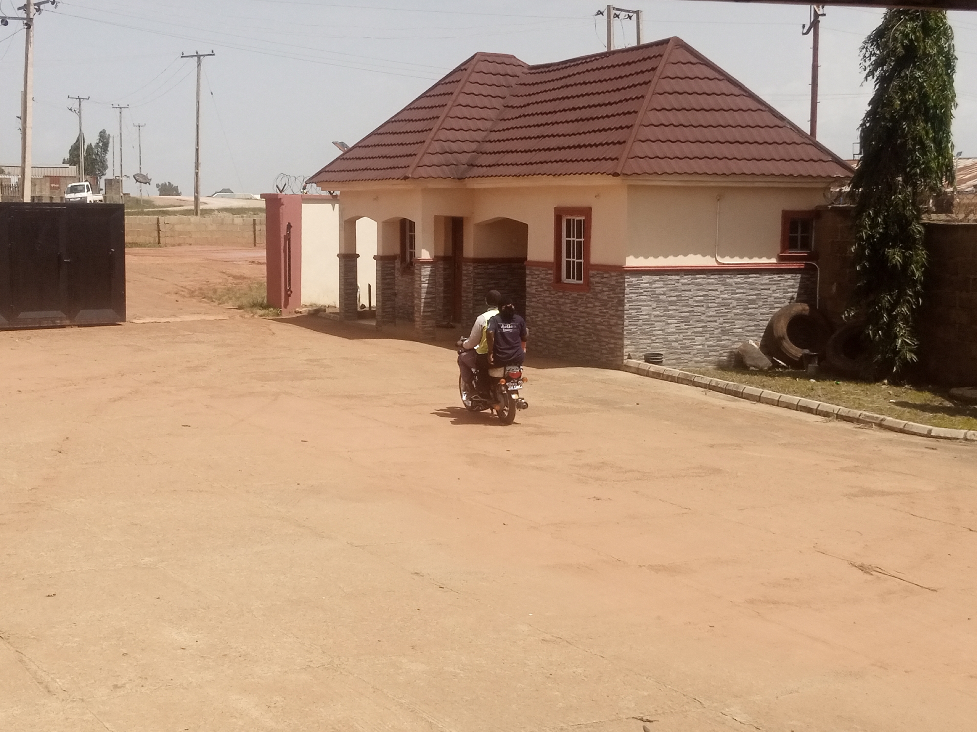 This media file depicts an outdoor scene with a building, a dirt road, and two people on a motorcycle. The building is a light-colored structure with a dark-pitched roof and decorative stone siding on its lower half. It has several windows, some with white frames and others with darker frames. To the left of the building, a dark metal gate is partially open. In the background, there are utility poles with wires, a white van, and a tall, leafy green tree.
The two individuals are riding on a motorcycle, with one person driving and the other seated behind them. They are facing away from the camera, moving across the dirt road. The rider in the back is wearing a dark blue shirt with white text that is partially obscured. The overall setting appears to be a somewhat dusty or undeveloped area, possibly a residential or commercial complex under construction or in a rural location. The sky is a pale, hazy blue, suggesting it might be a warm or slightly overcast day. There are no strong indicators of a specific time of day or strong emotions conveyed by the subjects. The scene is static, capturing a moment in time as the motorcycle passes by.