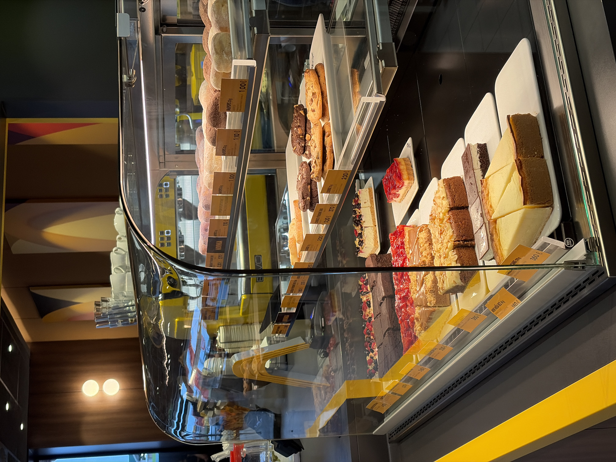 The image shows a display case filled with various desserts, including cakes, cookies, and pastries. The display case is well-lit, and the desserts are arranged neatly on shelves. In the background, a yellow coffee machine is visible, along with cups and other cafe equipment. The overall impression is that of a clean and modern cafe or bakery. This image was taken in a McDonald's in Nitra, Slovakia, which features a McCafe section.