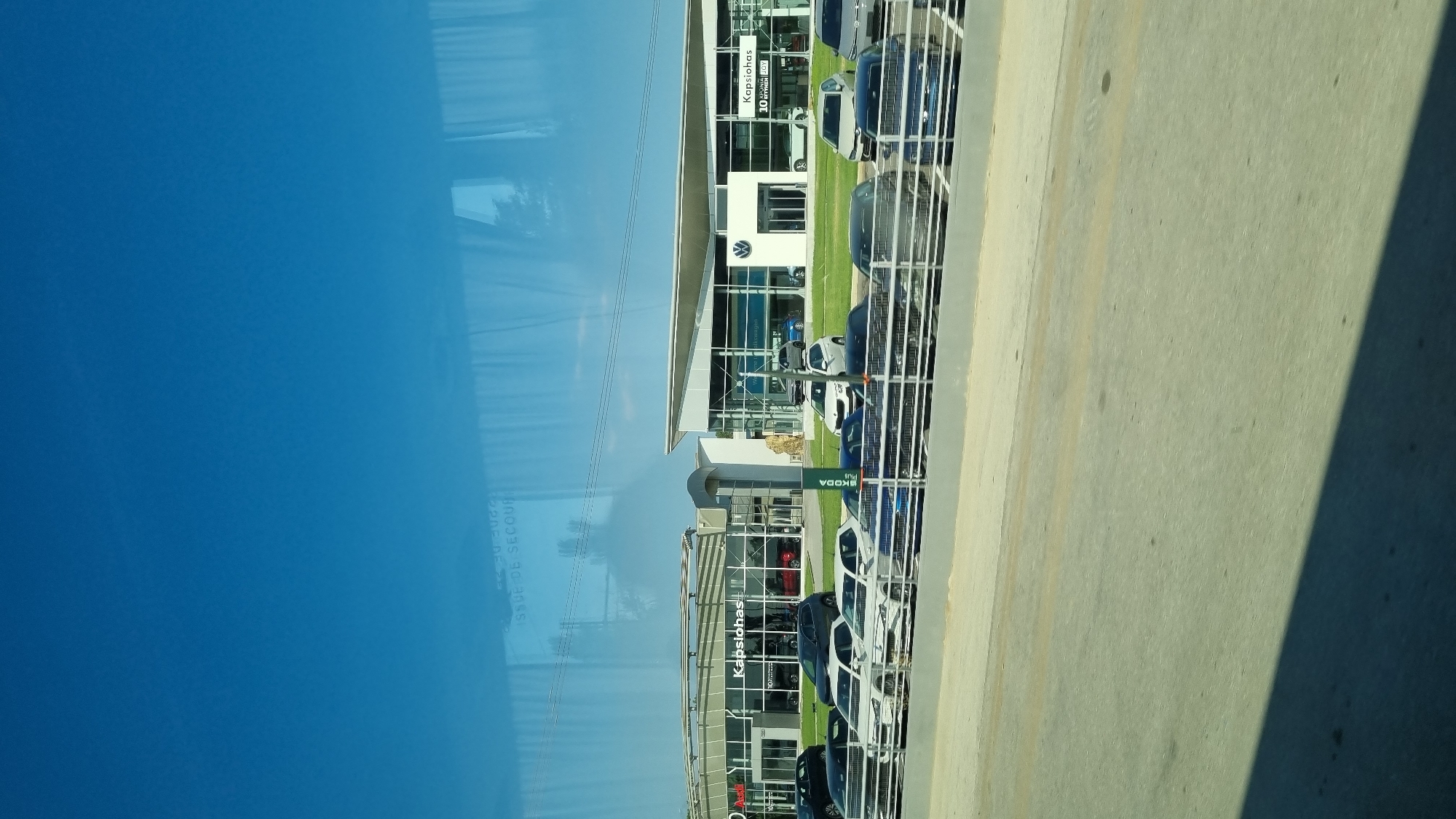 A vertical, eye-level shot shows a modern car dealership with a clear blue sky in the background. The dealership features large glass windows and a curved, contemporary roofline. Several cars are parked in front of the building, both on the lot and in a display area separated by a low metal fence. The dealership's signage includes "Kapsiotis" and a Volkswagen logo. Reflections of trees and buildings are visible in the glass of the dealership and the car windows. The foreground shows a paved road and sidewalk. The image is a photograph taken from a slight distance, possibly from another vehicle or a slightly elevated position. The overall scene suggests a commercial area on a bright, sunny day.