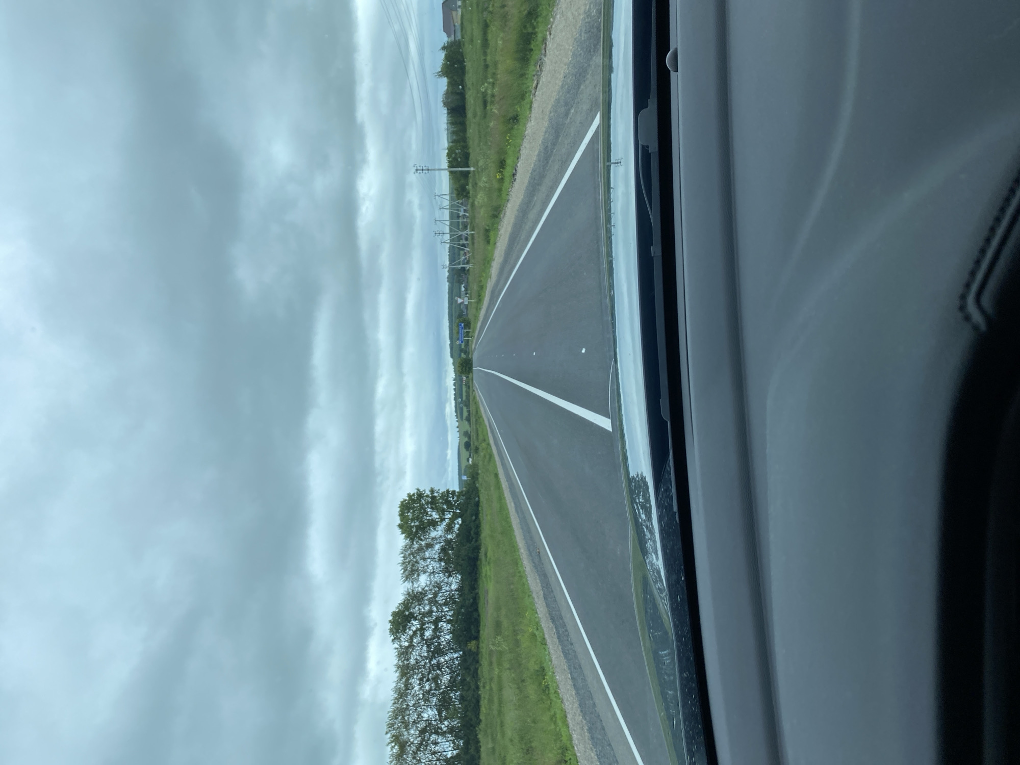 A driver's perspective shot captures a cloudy day on a two-lane asphalt road. The road has clear white lane markings and shoulders lined with gravel and grass. To the left, a line of trees runs along the edge of the road, beyond which lies a grassy field. To the right, power lines stretch across the landscape, with a few houses visible in the distance. The foreground shows the dashboard and windshield of a vehicle. The scene is set in Kliuchiki, Russia.