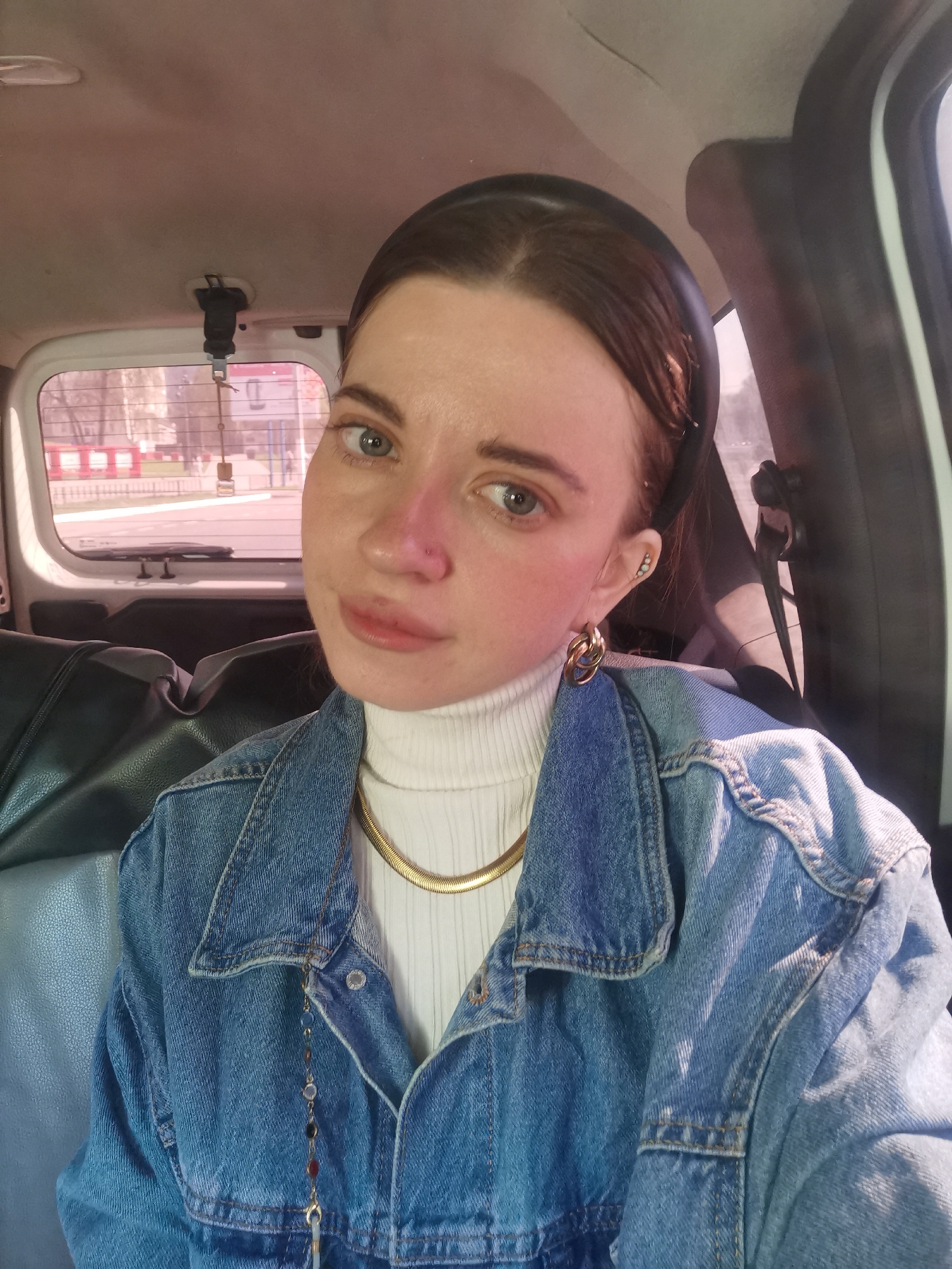A woman is seated inside a vehicle, likely a van, as suggested by the seating visible in the background. She is wearing a black headband, a white turtleneck sweater, a gold chain necklace, a denim jacket, and gold hoop earrings. She has a nose piercing and possibly additional ear piercings. The view through the back window shows buildings in the city of Chaikovsky, Russia, recognizable by the distinctive red and white barrier and surrounding architecture. The vehicle is parked or moving slowly on a road in an urban environment.