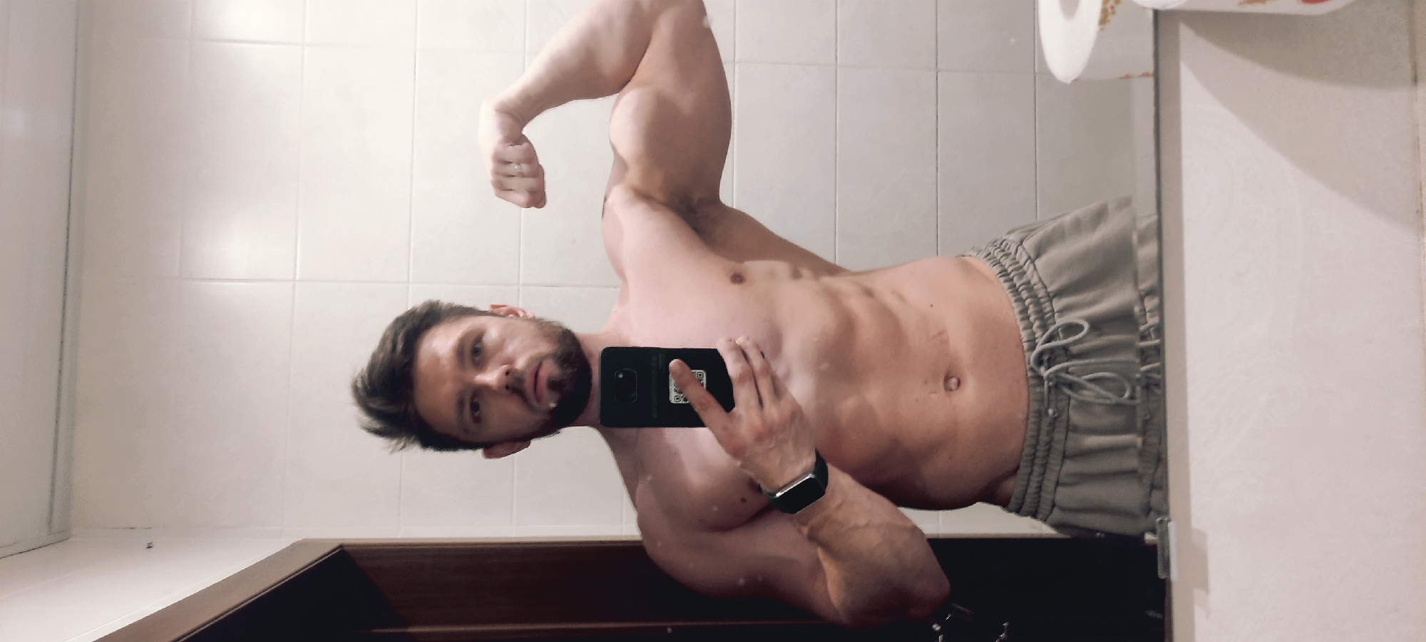 A man is captured in a mirror selfie, posing indoors. The image is oriented sideways, but depicts the man standing upright, flexing his left bicep in front of a mirror. He has a muscular physique, short dark hair, and a neatly trimmed beard. He is shirtless, revealing well-defined abdominal muscles, and wears light grey, drawstring sweatpants or shorts. A silver ring is visible on the ring finger of his right hand. He holds a black smartphone in his right hand, covering part of his face, and a black smartwatch is worn on his left wrist. The back of the smartphone shows a QR code. His expression is neutral.
The setting appears to be a bathroom or similar utility room, indicated by the white tiled wall visible behind him and reflected in the mirror. A roll of toilet paper on a holder is also visible in the upper right portion of the mirror's reflection. A dark wooden counter or cabinet is visible below the mirror. The lighting is artificial, suggesting an indoor environment, possibly in Fryazino, Russia. No specific time of day or weather conditions are discernible.