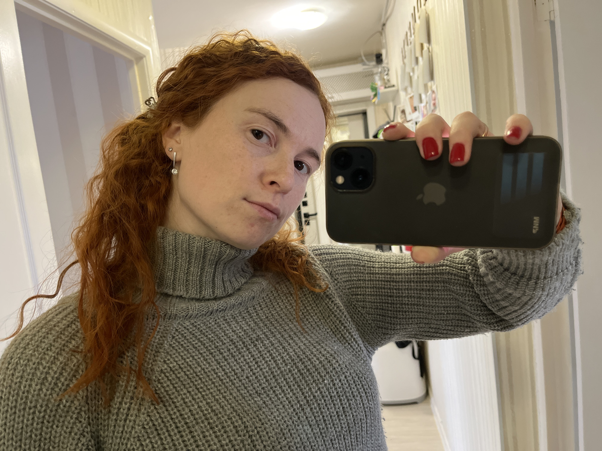 A woman with reddish-brown, curly hair, partially pulled back by a silver clip, is taking a selfie indoors. She has freckles on her face and a neutral expression, looking directly towards the camera. She wears a grey, chunky knit turtleneck sweater. In her right hand, she holds a dark-colored iPhone, identifiable by the Apple logo and triple camera module, encased in a dark protective cover that has the white text "KNM" visible. Her fingernails are painted bright red. Her left ear features two silver earrings: a small stud and a dangling one with a clear stone.
The setting appears to be an indoor hallway or room. On the left, a white door frame is visible. The background on the right shows a light-colored wall adorned with several small items, possibly photos or notes, and a white shelf or surface. Overhead, a circular ceiling light illuminates the scene. The lighting suggests an interior space during the day or well-lit by artificial means. The woman is the central focus, engaged in the act of self-portraiture.