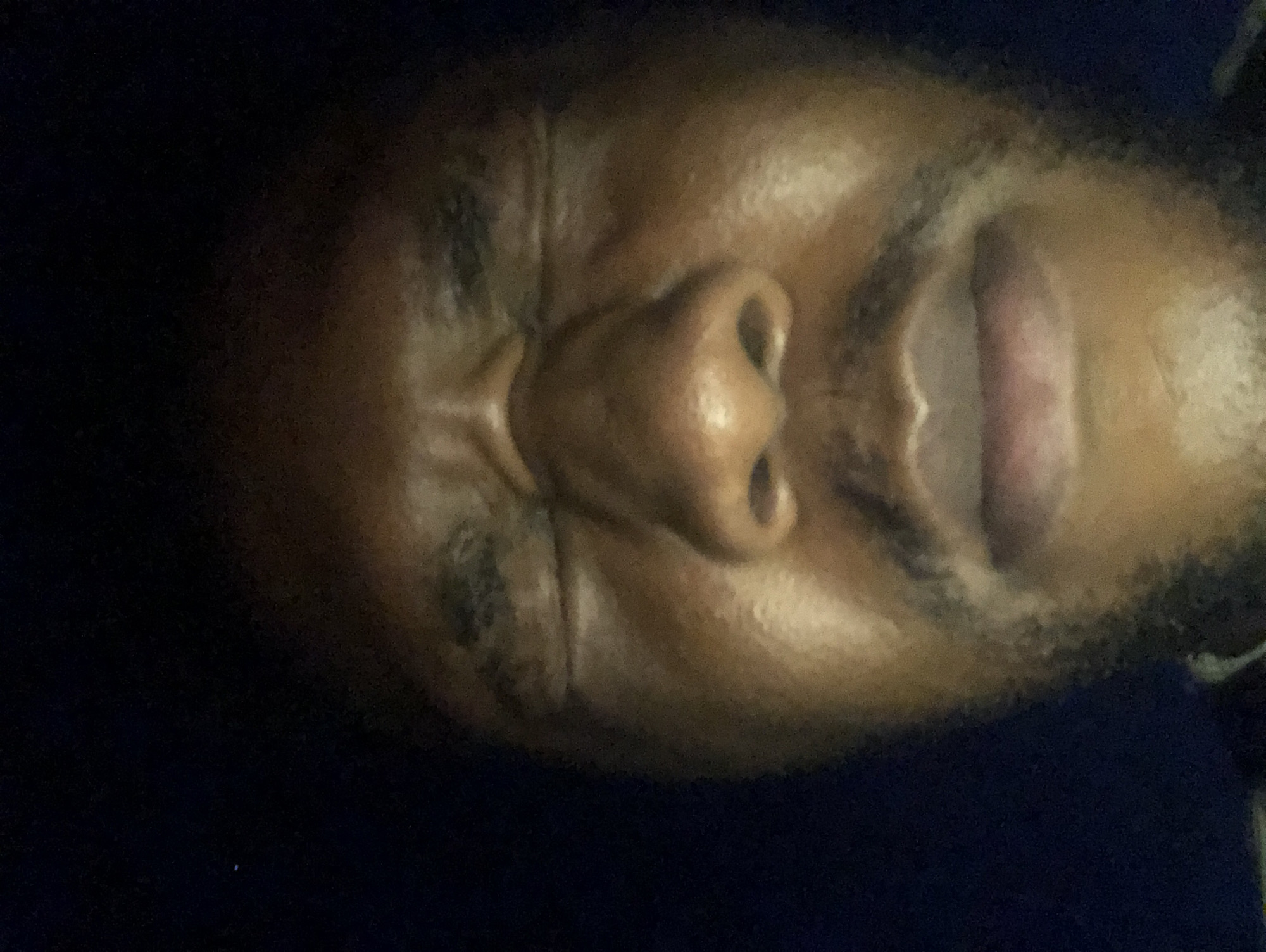 A close-up, tilted portrait shot shows a dark-skinned person with their mouth open. Features visible include the nose, lips, and facial skin. The background appears dark. The city and specific location are unidentifiable.