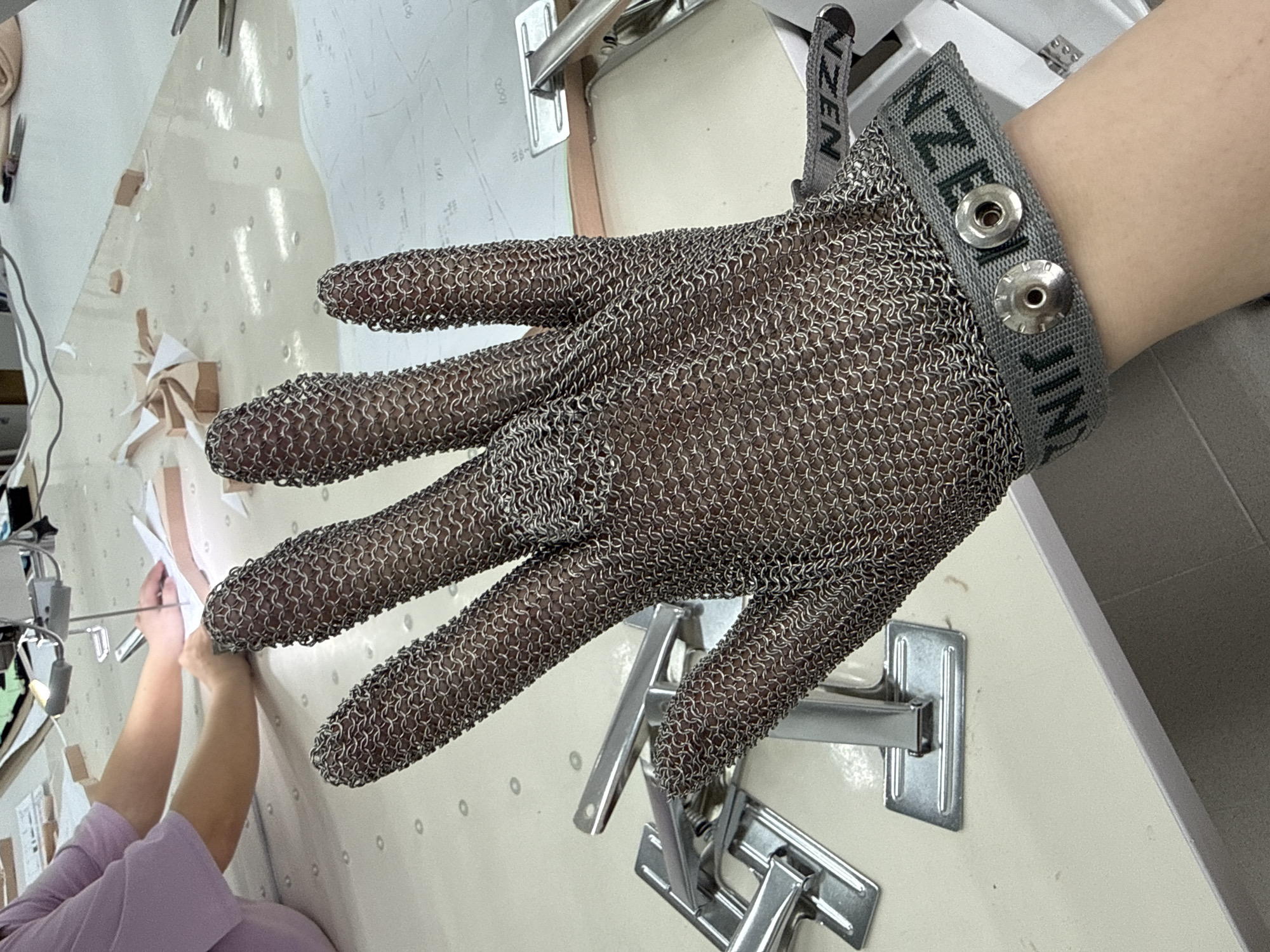 A person wearing a chainmail glove with a grey wrist strap featuring two metal snaps is visible. The brand name "NZEN JIN" is printed on the strap. The glove-wearing hand is extended and partially obscured by various pieces of metal hardware. The background consists of a light-colored surface with markings resembling patterns and a partial view of another person in a light purple shirt. The location cannot be determined from the image.