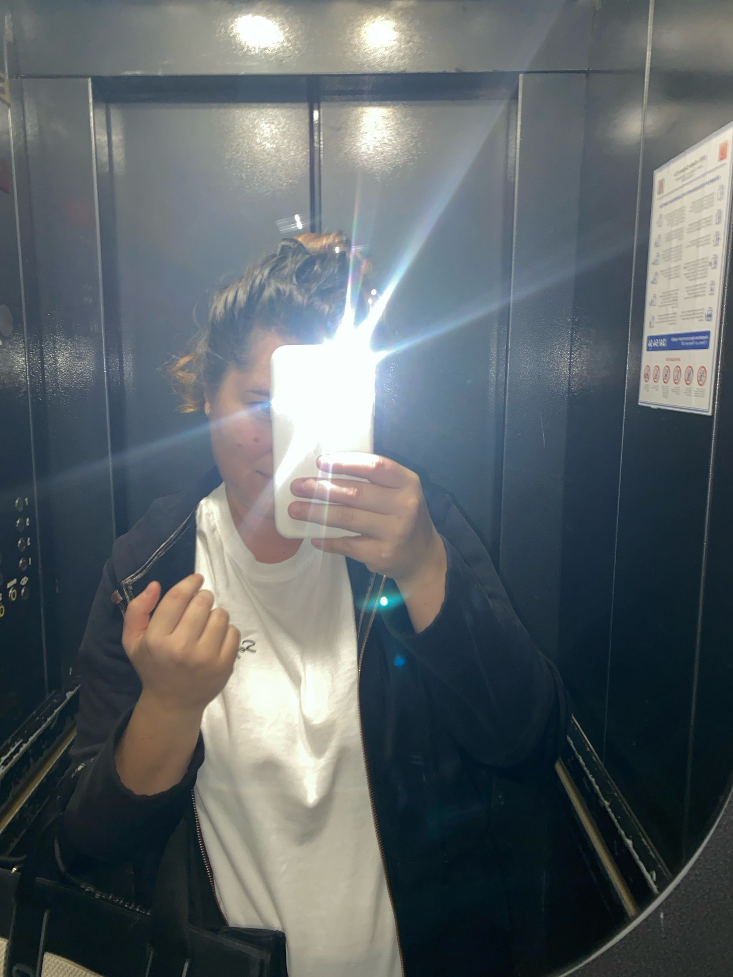 A young woman is taking a selfie in an elevator. She is holding a white smartphone in her right hand, which is reflecting light directly into the camera, obscuring her face. She is wearing a white t-shirt and a black zip-up jacket. Her hair is pulled up in a messy bun. The elevator walls are dark gray, with some buttons visible on the left. A safety notice is posted on the right wall. The scene appears to be indoors, possibly in an apartment building or office. The city of Perm, Russia is mentioned in the prompt, but there are no visual cues in the image to confirm this specific location.