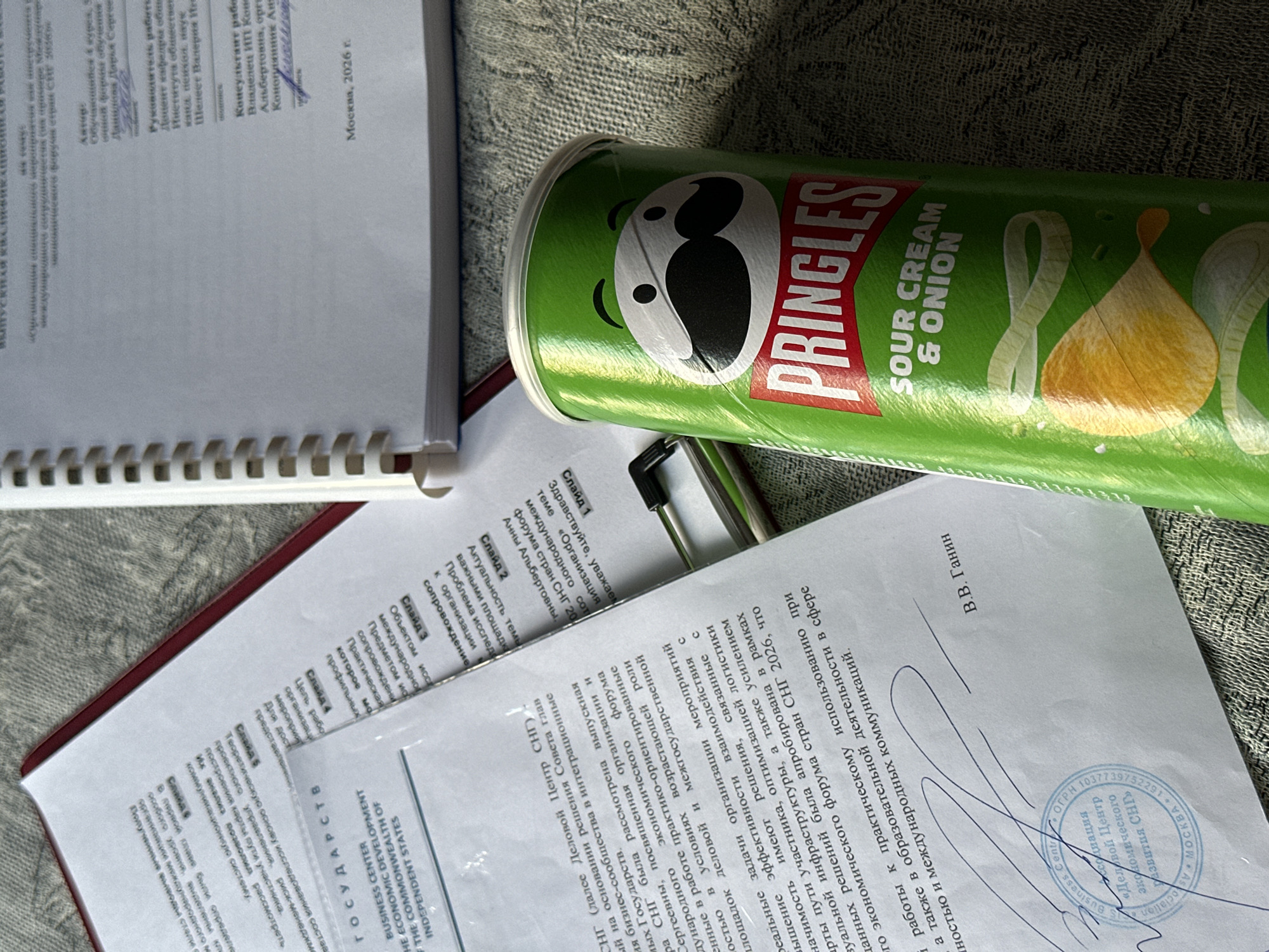 The image shows a close-up view of a Pringles can, specifically the Sour Cream & Onion flavor, placed on a textured surface, likely a fabric. To the left of the can, there are several documents, one of which is a white paper with Russian text and the logo of the "BUSINESS CENTER OF THE ECONOMIC DEVELOPMENT OF THE COMMONWEALTH OF INDEPENDENT STATES". This document also has a stamp with "MOCKBA" (Moscow) and "2026 r." (year 2026) indicating a connection to Moscow, Russia, and a future date. Another document with a red binding and Russian text is partially visible. A spiral-bound notebook with white pages is on the right side of the image. The lighting suggests an indoor setting. No people are visible in the image.