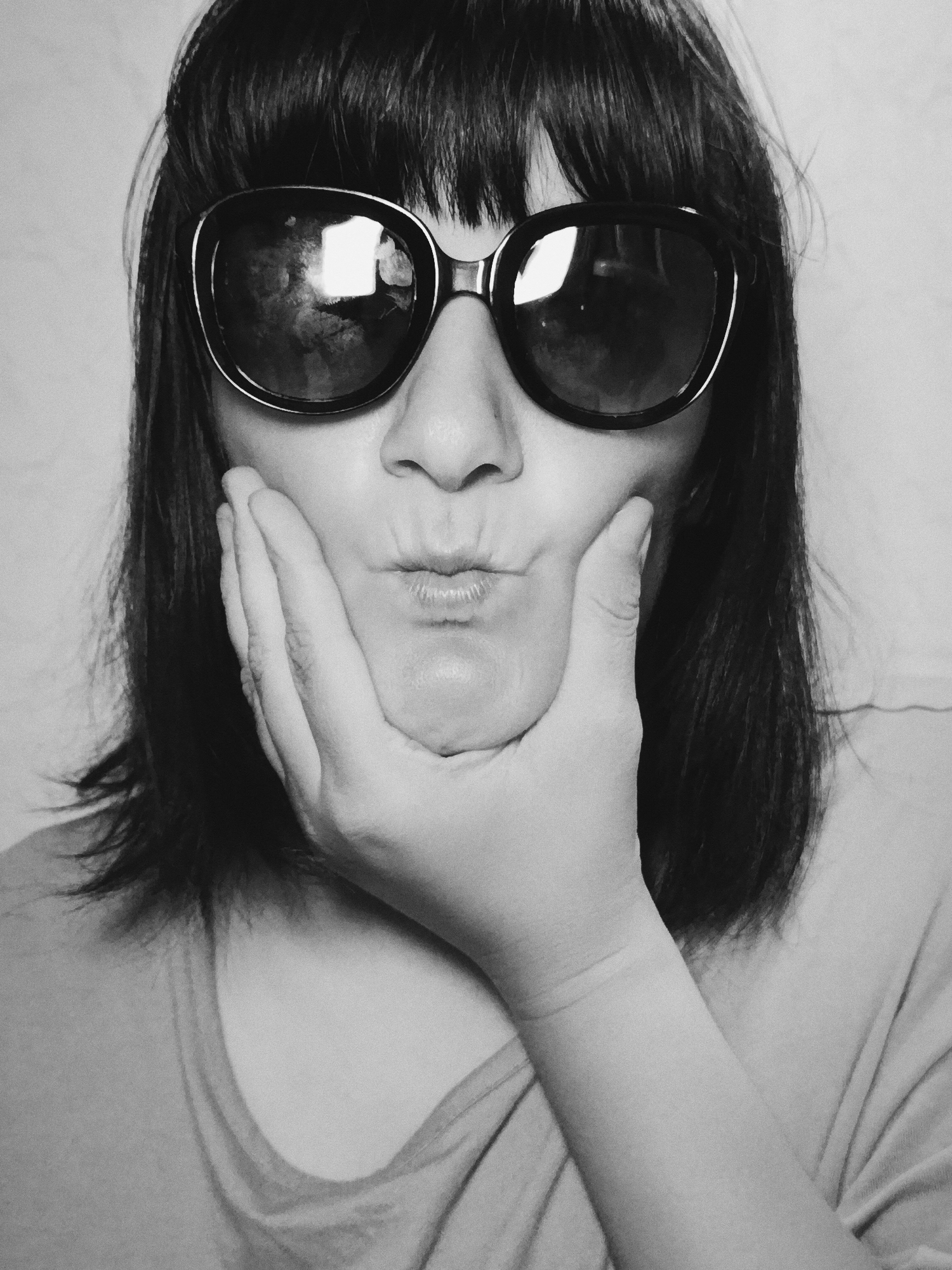A monochrome, close-up portrait features a person with dark, straight hair and bangs. They are wearing large, dark sunglasses that obscure their eyes. The person's hand is positioned against their cheek, supporting the face, and their lips are pursed in a pouting or kissing expression. The background is a plain, light surface. No location details are discernible.