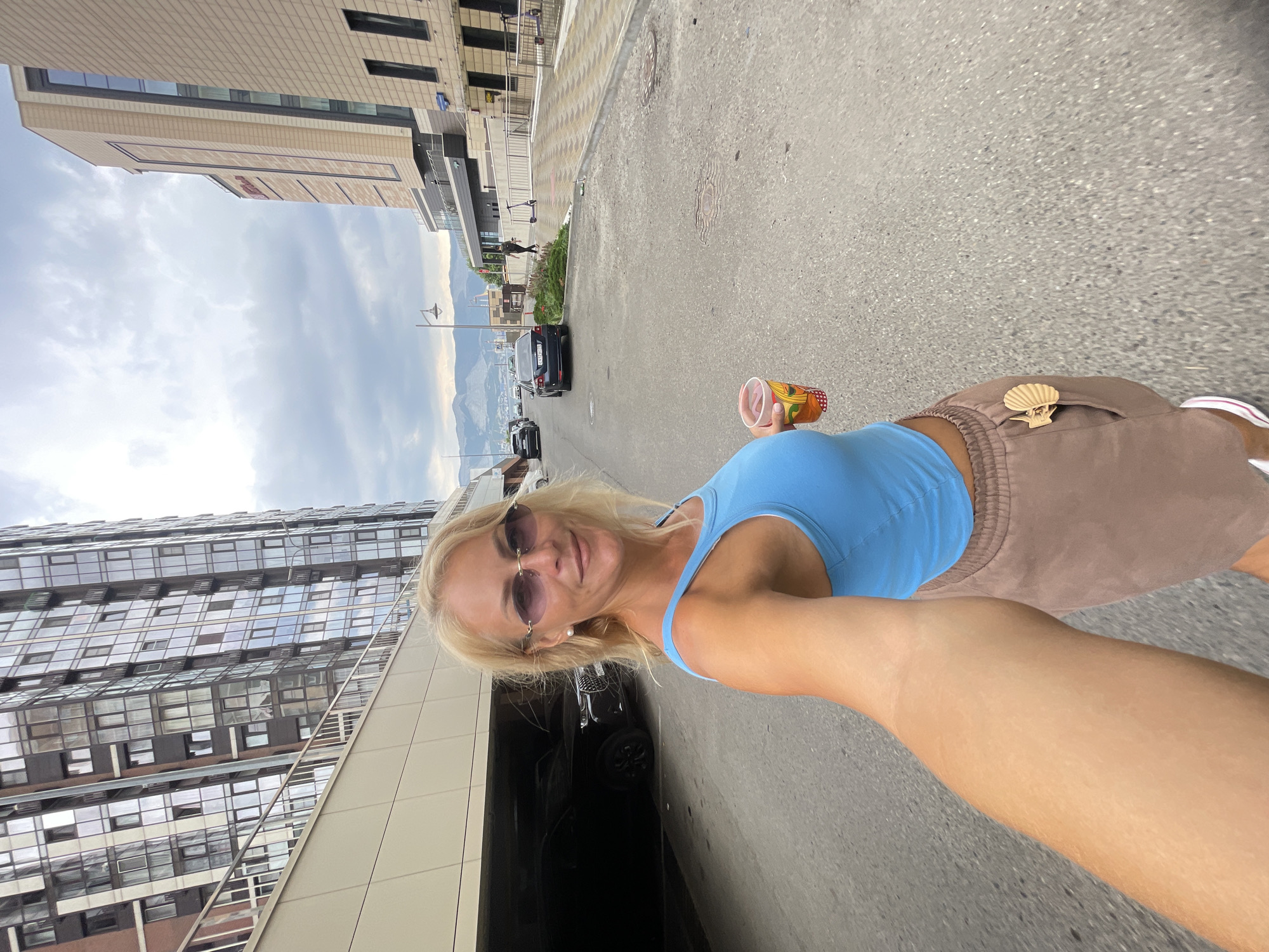 A blonde woman in a blue crop top and brown shorts holds a drink in her right hand, taking a selfie in an urban environment. She wears oval-shaped sunglasses and a pearl earring. Behind her, a multi-story building with reflective windows dominates the left side of the frame, partially obscuring a covered parking area with dark cars. To the right, a light-colored building with a glass facade extends into the background, with a street running alongside it. The sky is overcast with clouds, and mountains are visible in the distance. The ground is paved asphalt.