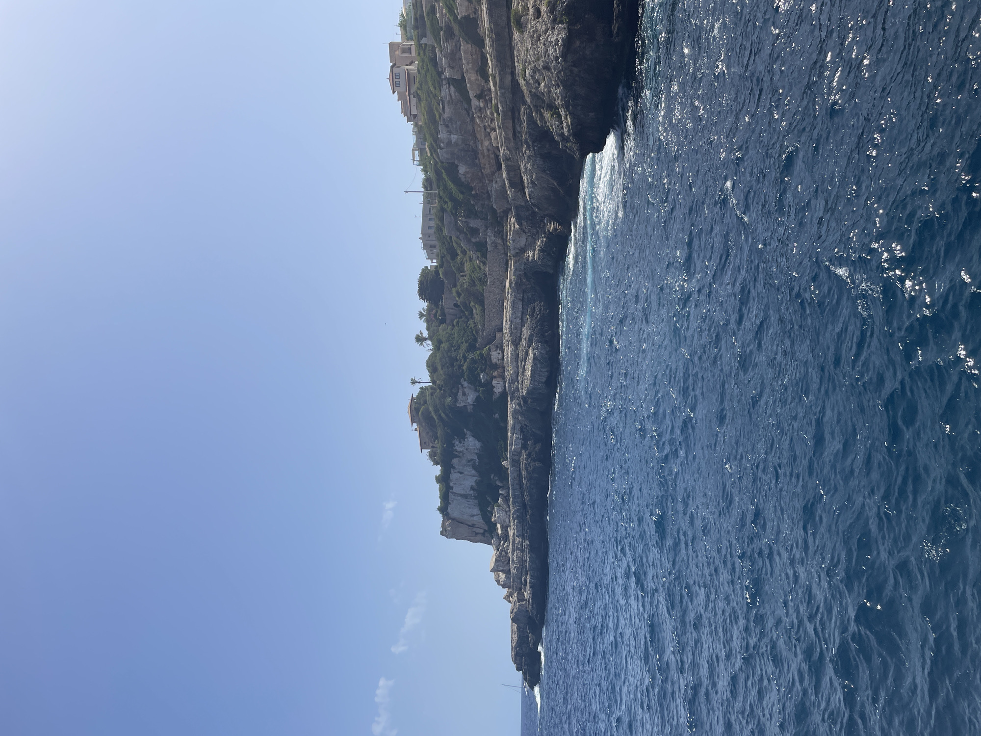 An eye-level, medium shot captures a bright, sunny day with a clear blue sky and a vast expanse of deep blue ocean. The water exhibits gentle ripples and sparkles from the sunlight. To the right, a rugged, rocky coastline rises, topped with lush green vegetation. Several buildings, possibly villas or houses, are visible nestled among the greenery on the cliff tops. The architecture appears to be Mediterranean-style, with tiled roofs. A small sailboat with a white sail is present in the distance on the left side of the frame, navigating the ocean. The overall scene depicts a scenic coastal landscape in the vicinity of Santanyí, Spain.