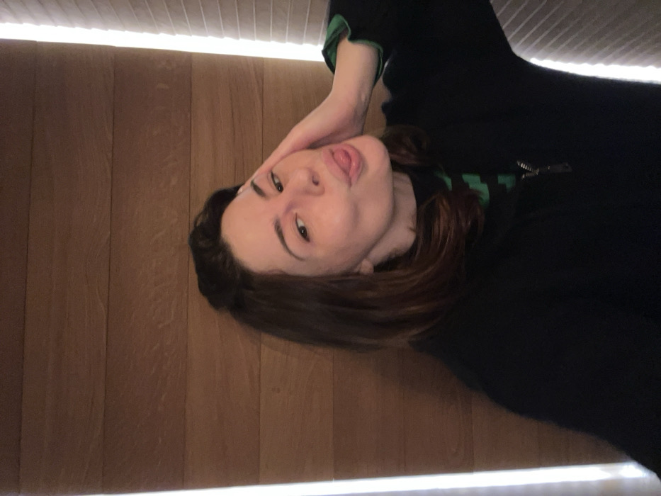 A woman with dark hair and light skin poses with her head tilted, sticking her tongue out slightly. She wears a dark, long-sleeved top with green accents. Her left hand is gently touching her face near her cheek. The background features light wood paneling, with a bright light strip running along the top and bottom of the image, creating a border. The photo is taken at an unusual angle, rotated counterclockwise.
