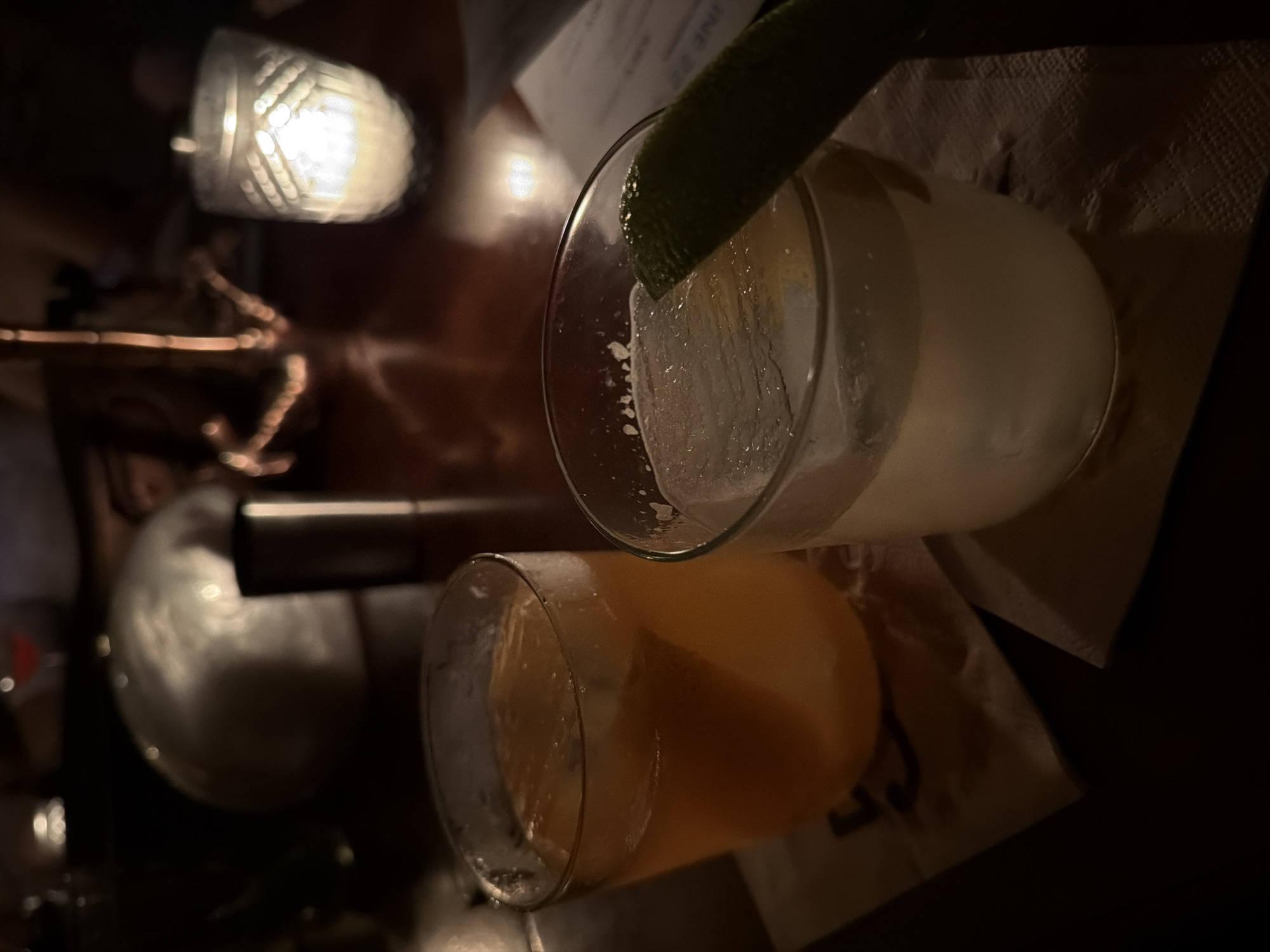 The image captures a low-lit indoor setting, possibly a bar or restaurant, with two partially consumed cocktails as the focal point. One cocktail is an amber color, the other a lighter color and garnished with a lime wedge. Both are resting on napkins with visible text and logos. To the left of the cocktails, a blurred metal object, possibly bar equipment, is visible. Above that, a light fixture with a patterned glass shade is lit. The location of the scene is unspecified but identified in the prompt as Sofia, Bulgaria. No people are clearly visible.