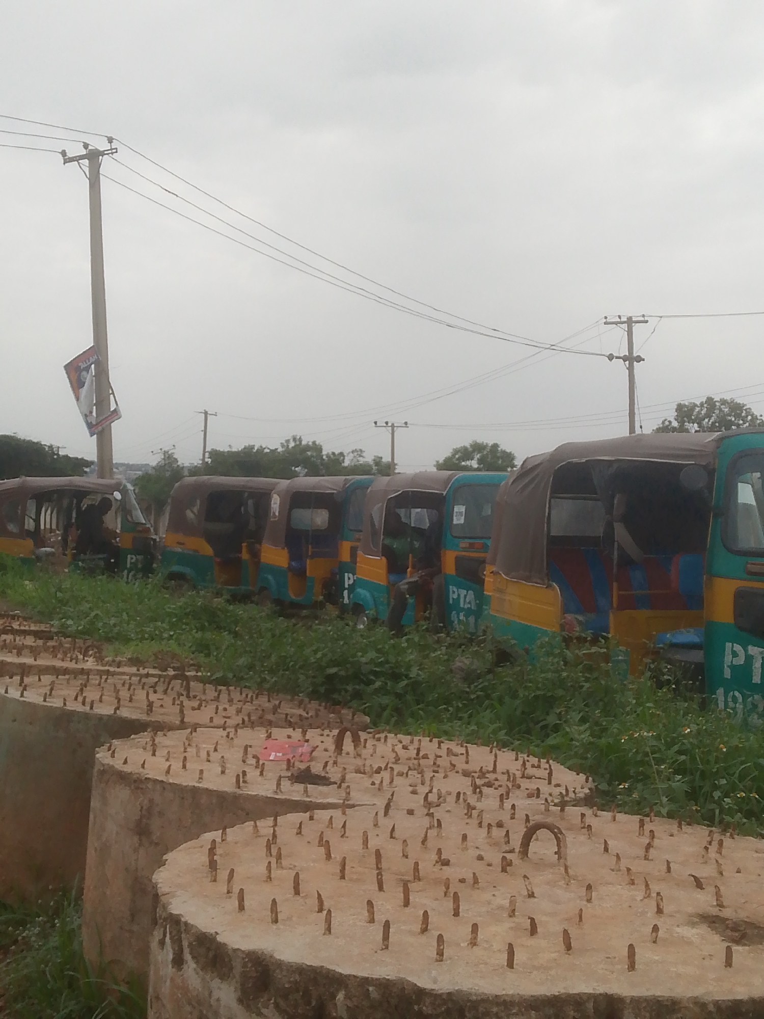 In Jos, Nigeria, a line of parked auto-rickshaws, commonly known as "Keke NAPEPs," are visible under an overcast sky. The vehicles, predominantly colored in teal and yellow, are parked nose-to-tail in a grassy area. Some of the Keke NAPEPs have tarpaulins covering their passenger areas. A utility pole with electrical wires runs diagonally across the upper portion of the frame, and a poster is attached to the pole. In the foreground, a series of large, circular concrete structures, possibly for construction or infrastructure, are laid out, each studded with numerous metal rods. The overall scene suggests an outdoor storage or staging area for vehicles and construction materials, set against a backdrop of a cloudy sky and distant trees.