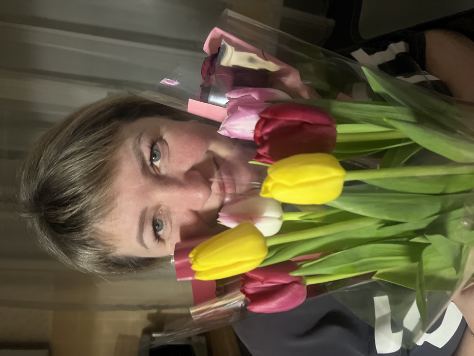 An adult female, identified as being in Chaikovsky, Russia, is depicted from the chest up, holding a bouquet of mixed-color tulips. She has fair skin, blue eyes, and short, light brown hair styled with bangs. Her expression is a direct smile towards the camera. She is wearing a dark, possibly charcoal-colored top. The bouquet consists of at least seven tulips, including prominent yellow, dark red/magenta, and light pink/white flowers, wrapped in clear plastic film with pink accents. The flowers obscure the lower portion of her face and torso. The background is an interior residential setting, dimly lit and out of focus, revealing a light-colored appliance on the left and a textured wall or curtain with vertical lines behind her.