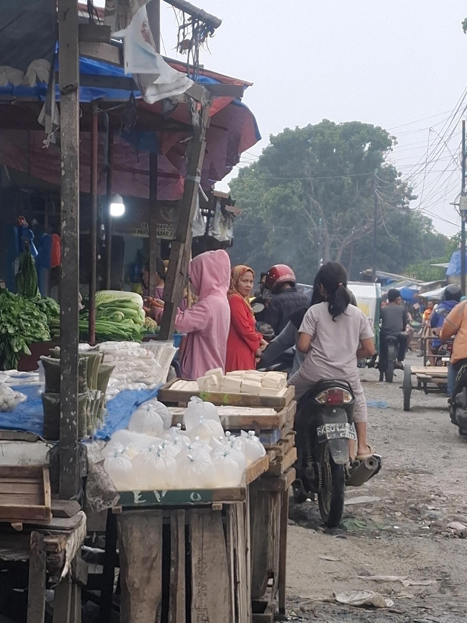 The image depicts a bustling market scene in Kenangan, Indonesia. In the foreground, a stall displays an assortment of fresh produce, including large heads of leafy green vegetables and packages of white food items, possibly tofu or tempeh, wrapped in clear plastic. A person wearing a pink hooded jacket stands near the produce, looking towards the stall. Behind this stall, another vendor is visible with more vegetables.
The middle ground reveals several people and vehicles. A woman in a red outfit and headscarf is talking to someone on a motorcycle. Two individuals are riding on a motorcycle, one driving and another as a passenger, both facing away from the camera. Further back, other market-goers and motorcycles are present, suggesting a busy commercial area. Power lines and utility poles are visible in the background, along with trees and the overcast sky, indicating an outdoor setting. The ground is rough and uneven, characteristic of a local market. The overall atmosphere is one of everyday commerce and activity.