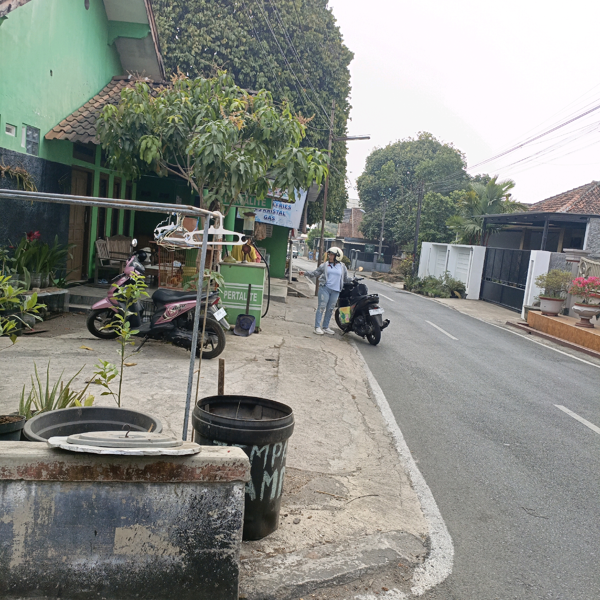This image captures a street scene in Salatiga, Indonesia, near a small roadside gas station. A woman wearing a white helmet, blue shirt, and ripped jeans stands on the sidewalk next to a black scooter, gesturing with her right hand. Beside her, a pink scooter is parked. The gas station has a green structure with visible signage that appears to advertise "PERTALITE" and "KRISTAL GAS". Behind the gas station, a two-story building with a green facade and a tiled roof is visible, with a tree in front of it. The street itself is paved, with a white line marking the edge of the lane. In the foreground, there are large planters and a dark gray bin with writing on it. The sky is overcast, and the environment suggests a moderately urban or suburban setting with trees and residential buildings lining the street.