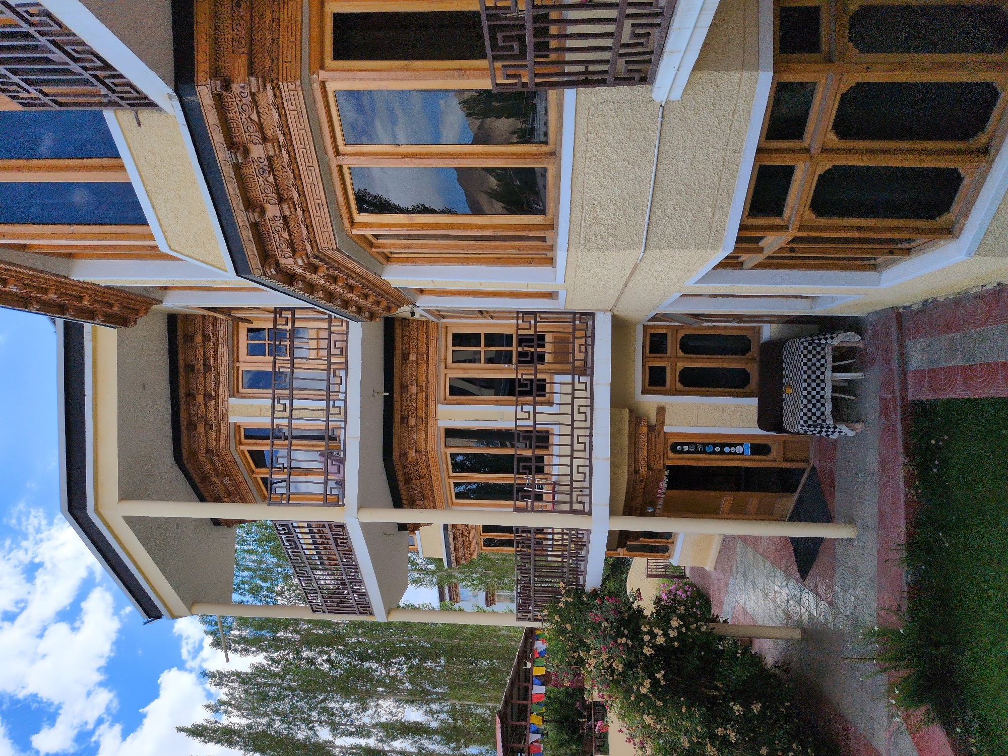 An exterior, low-angle shot captures a multi-story building with cream-colored walls and ornate wooden trim around the windows and balconies. The building features multiple balconies with dark metal railings and several windows, some reflecting the sky and surrounding landscape of trees and mountains. In the foreground, a path made of decorative tiles leads towards an entrance with wooden doors. To the left of the path, a lush bush with pink and white flowers is visible. A small table with a black and white checkered tablecloth is set up near the entrance. The scene is set against a backdrop of tall trees and a blue sky with white clouds. The overall impression is of a guesthouse or hotel, likely located in a mountainous region.