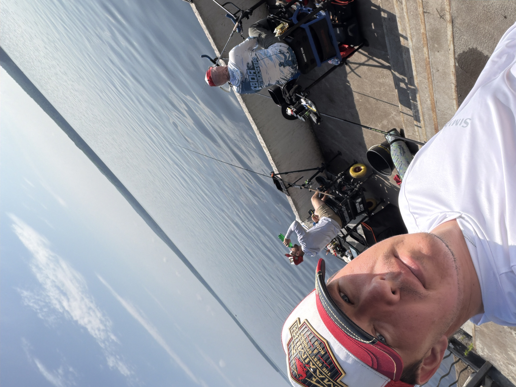 Three men are fishing from a concrete structure next to a large body of water, likely the Votkinsk Reservoir, near Chaikovsky, Russia. The man in the foreground is taking a selfie and wearing a white and red fishing hat. In the middle ground, a man in a white hoodie and khaki shorts drinks from a green bottle while fishing. Further back, another man in a white hooded shirt with Russian text and a red bandana is also fishing. Fishing rods are set up, and equipment such as buckets and a fishing chair are visible. The sky is blue with scattered clouds, and the water is calm with ripples. The sun appears to be shining, casting shadows on the concrete.