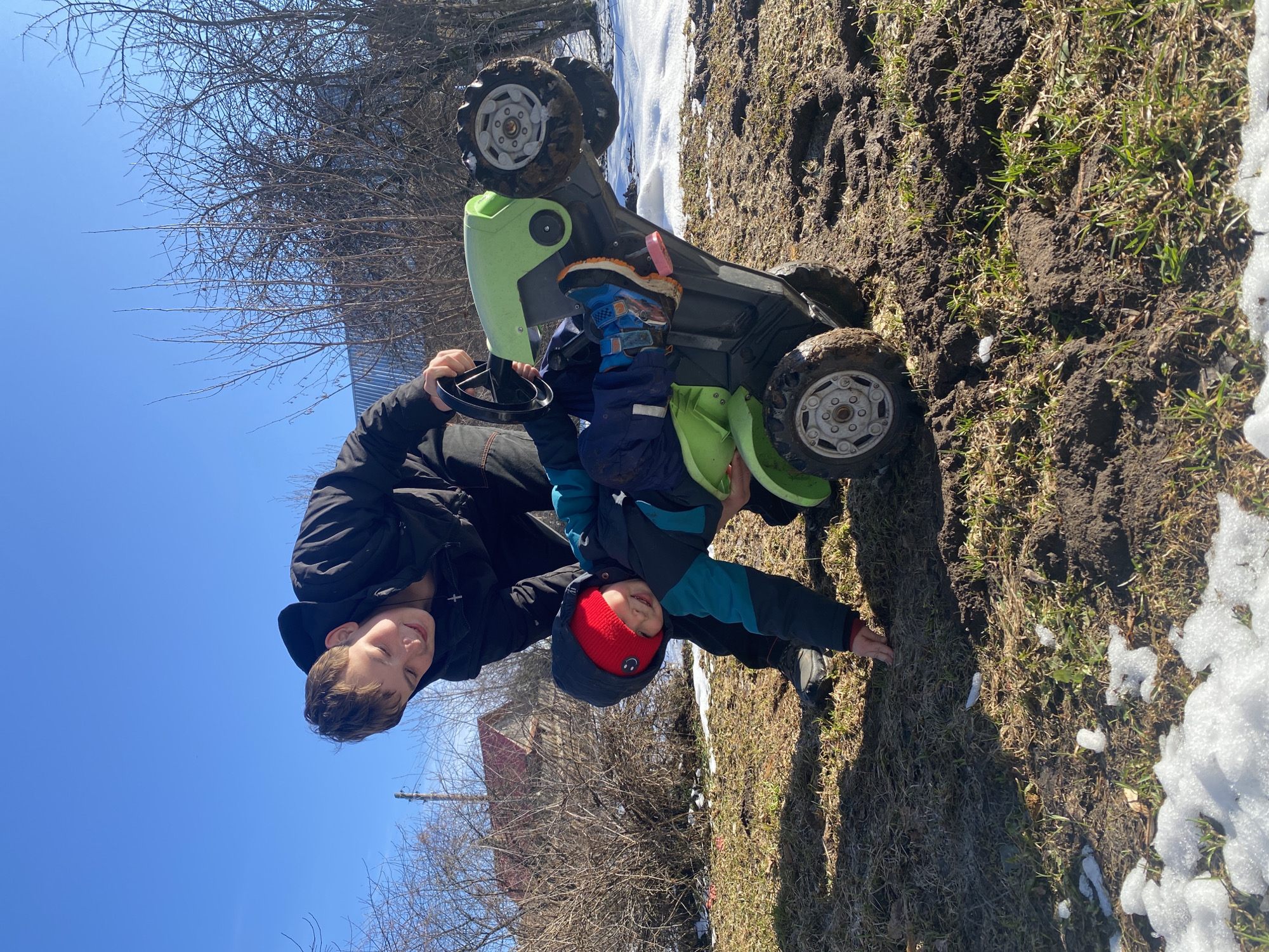 The image shows two boys and a toy car in an outdoor setting. The larger boy, wearing a black jacket, holds the steering wheel of a green and black toy car, while the smaller boy, wearing a blue jacket and red hat, is sitting in the car. The car appears to be overturned, with its wheels facing upwards. The ground is a mix of mud, grass, and patches of snow. In the background, there are bare trees, a building with a red roof, and a clear blue sky. The location is identified as Ольховка, Russia.