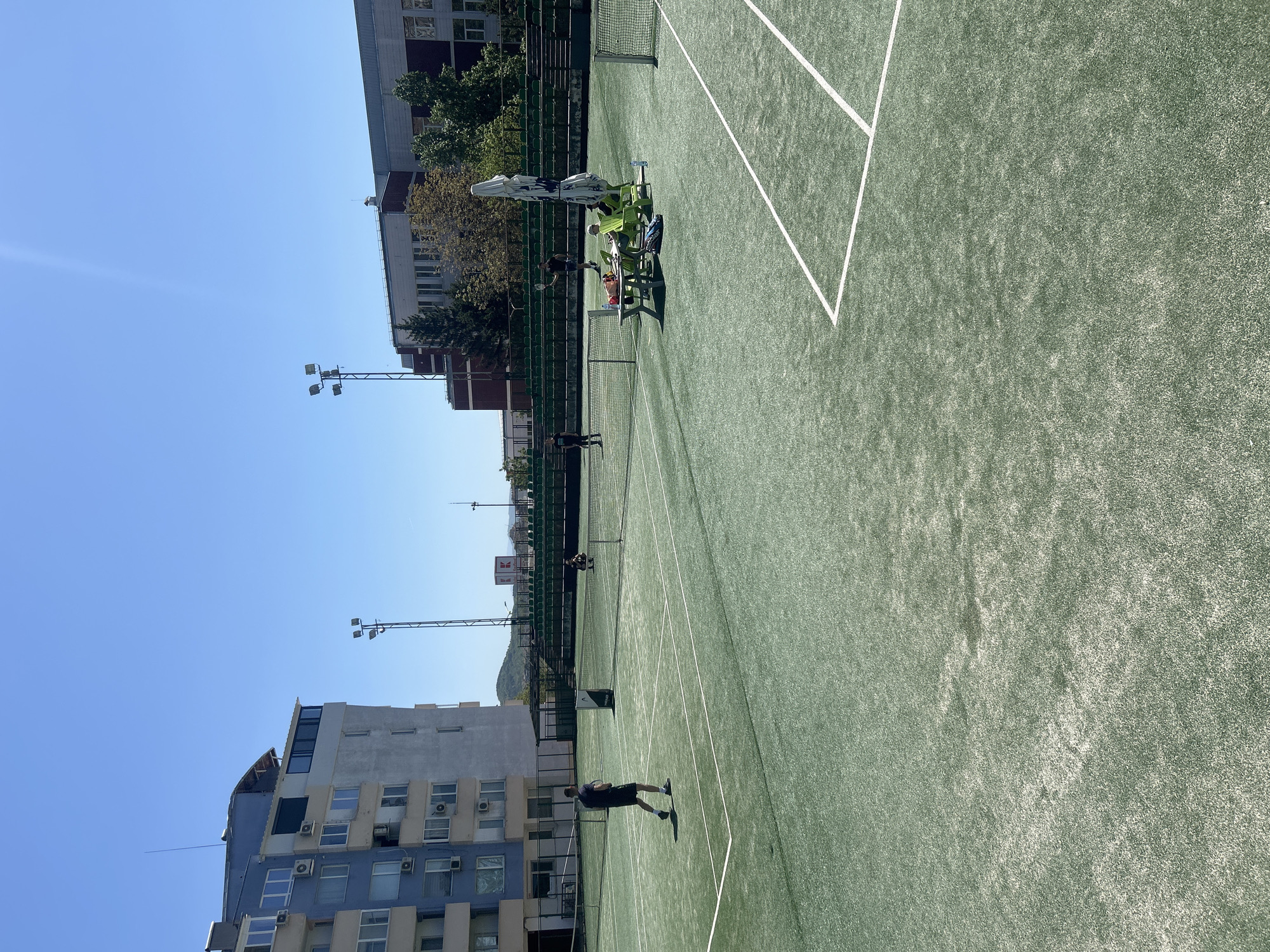 The image captures a tennis court in Veliko Tarnovo, Bulgaria, adjacent to urban buildings and under a clear blue sky. The tennis court's surface is green with white boundary lines. Several individuals are present on the court, with one appearing to be in motion, possibly playing tennis. Spectators are seated on chairs beside the court, shaded by an umbrella. Lighting towers are positioned alongside the court. The background shows buildings with a mixture of residential and commercial architecture typical of an urban environment.