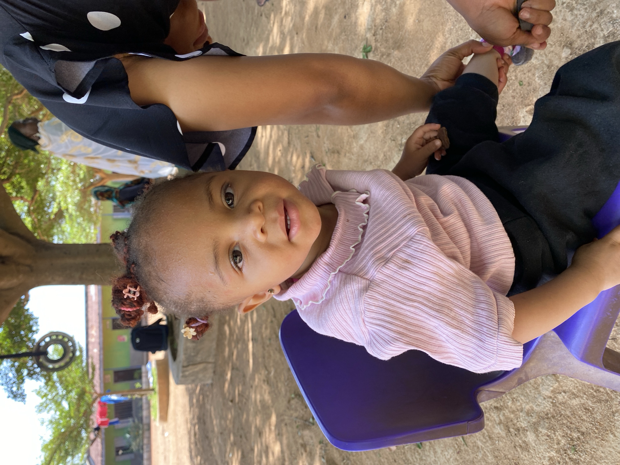 A young child sits on a purple plastic chair outdoors, wearing a light pink, ribbed, long-sleeved shirt and black pants. An adult, partially visible in a black and white polka dot dress, is holding the child's foot and attempting to put on a sock. The location is Gura Top, Nigeria, as indicated in the image details. The background shows trees, a building with a green exterior, and other indistinct structures. The ground is mostly dirt. The child looks at the camera. Small hair clips are visible in the child's hair.