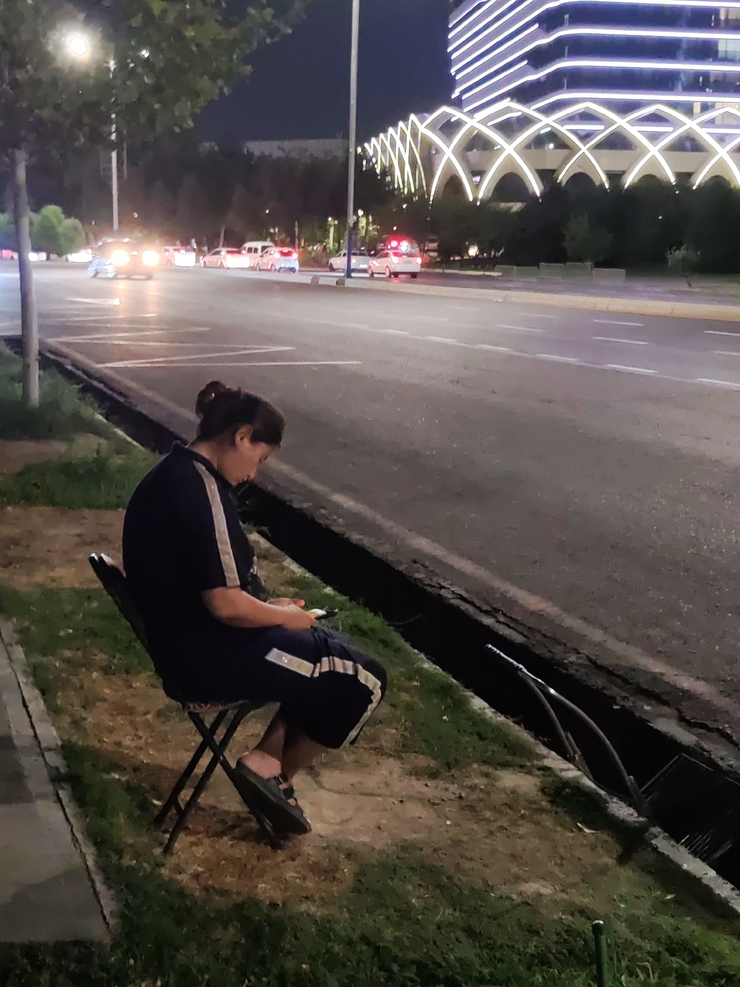A woman is sitting on a foldable chair on a grassy area next to a road at night. She is wearing a dark blue t-shirt with white stripes on the sleeves and dark blue shorts with white stripes. She is holding a mobile phone in her hands and looking down at it. The road is illuminated by streetlights and car headlights. In the background, there is a modern building with illuminated white lines and arches. There are cars driving on the road, and one car has its hazard lights on. The scene appears to be in a well-lit urban area of Tashkent, Uzbekistan, likely a main thoroughfare with pedestrian access.
