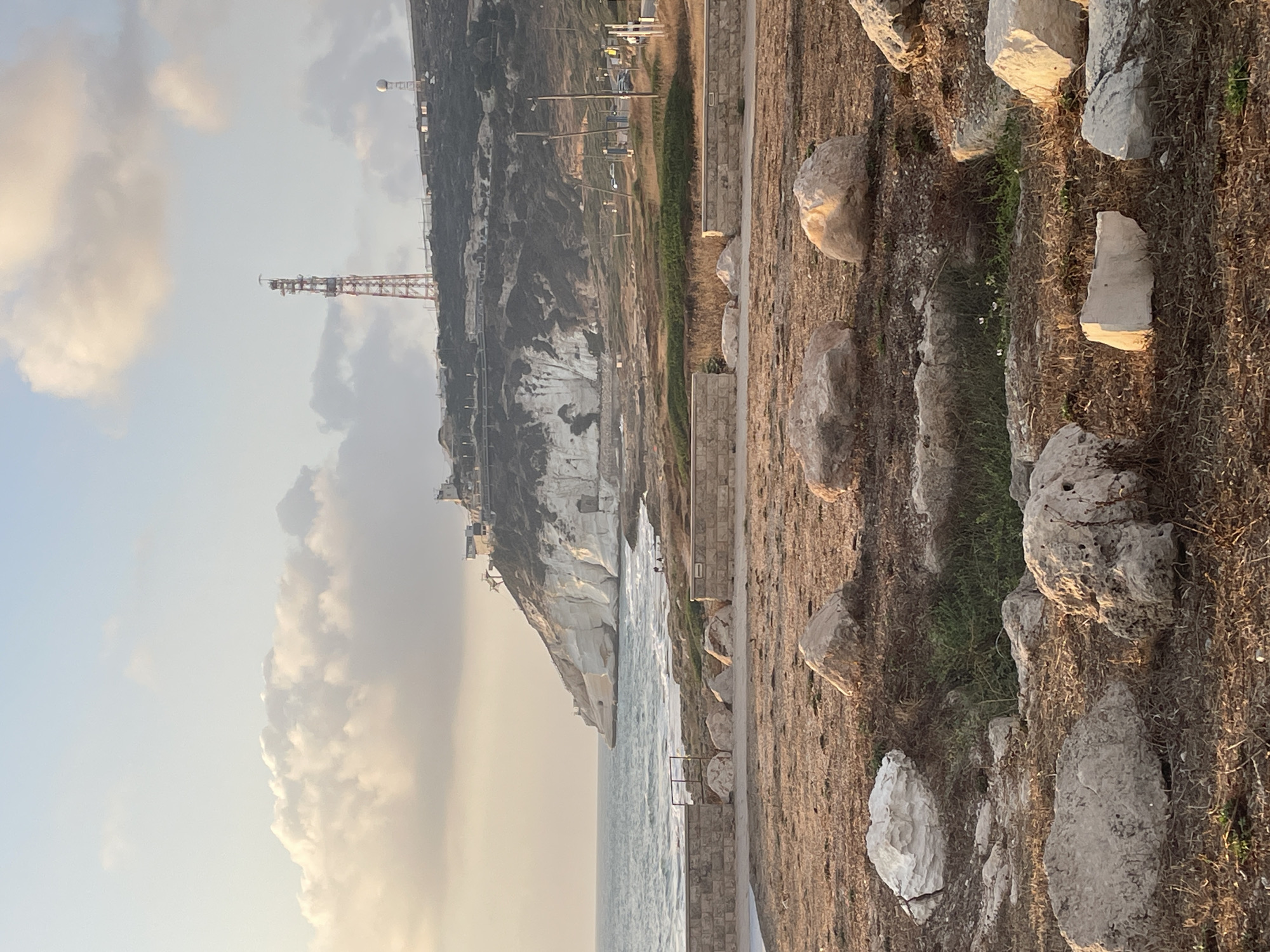 The image depicts a coastal landscape with a prominent white cliff face on the right side, overlooking the sea. A cell tower, red and white, stands atop the cliff, alongside other structures. The foreground is a dry, grassy area dotted with large rocks. In the distance, waves crash against the shore. The sky is partly cloudy with a pale, warm light suggesting either sunrise or sunset. The scene appears to be located in the city of Mate Asher Regional Council, Israel, given the characteristic coastal cliffs and vegetation. There are no people clearly visible in the image.