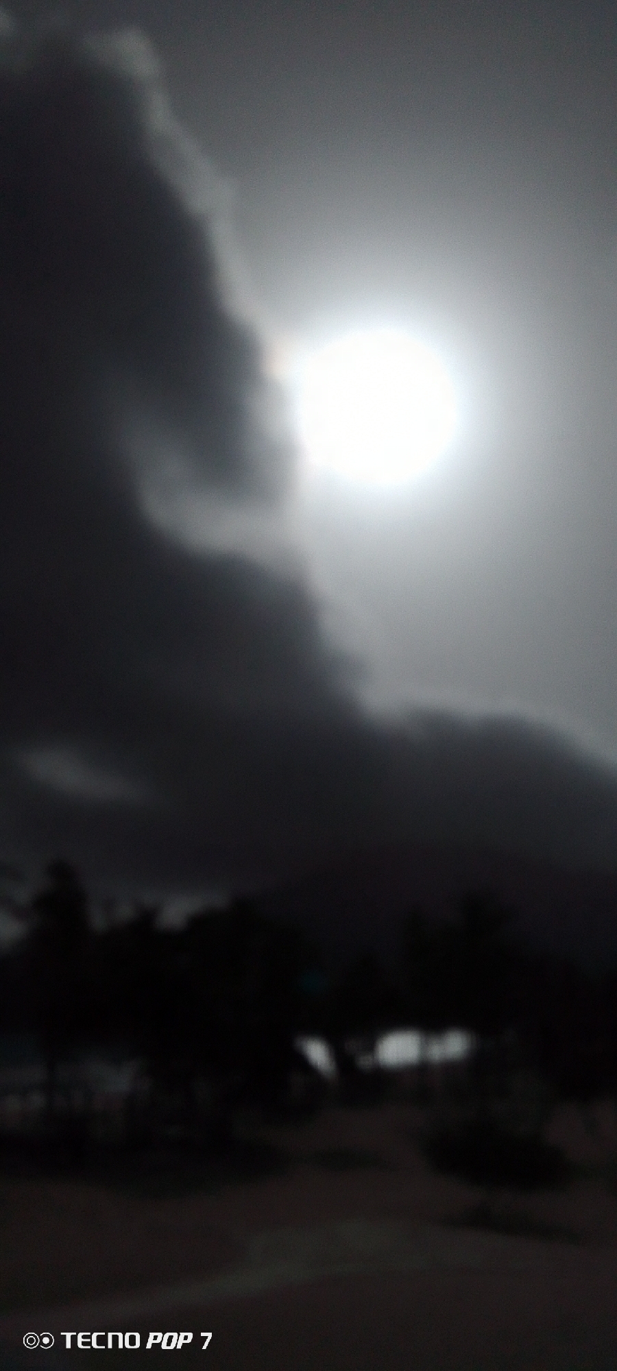 The image shows a bright, diffused sun behind dark, heavy clouds. The bottom of the image is dark and blurry, possibly showing trees or foliage in silhouette. The scene is dimly lit and appears to be overcast. "TECNO POP 7" is visible in the bottom left corner of the frame. The location in the city of Pedro González, Venezuela is not discernible from the image.