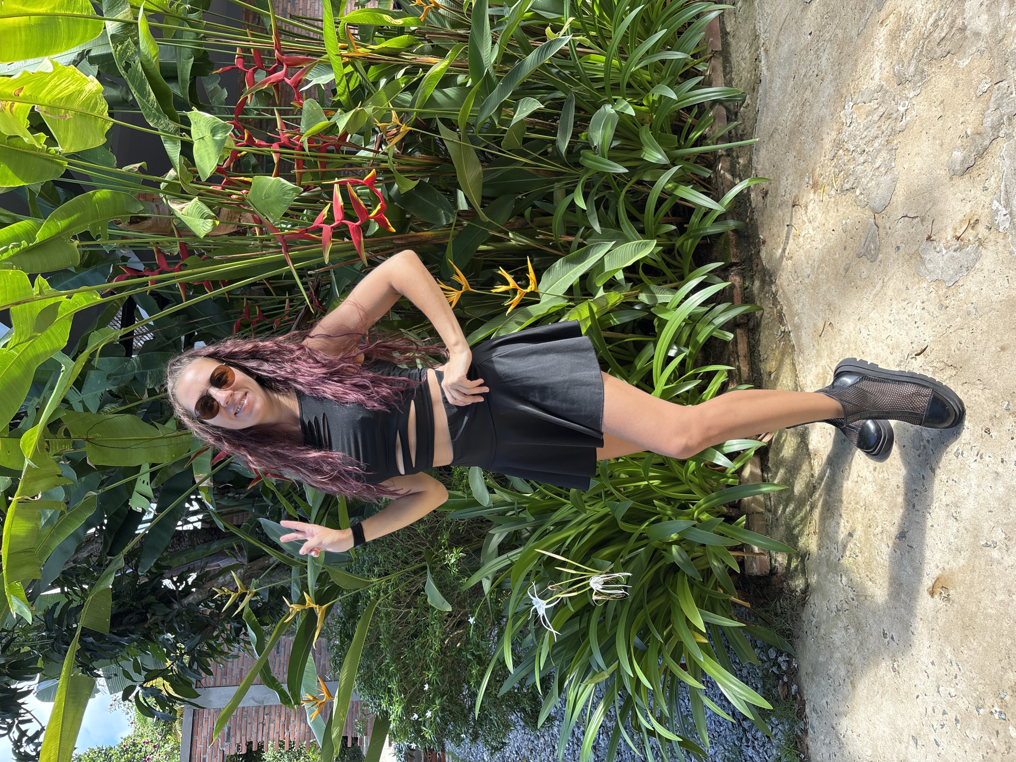 A young woman stands in front of a lush green foliage backdrop with red and yellow flowers. She is wearing a black crop top and a black mini skirt, paired with black mesh boots. Her long, wavy hair is purple and she has sunglasses on. She is posing with her left hand on her hip and her right hand making a peace sign. The ground is concrete with some gravel and a few small plants. The overall setting appears to be outdoors, possibly in a tropical or subtropical climate, with a brick wall visible in the background.
