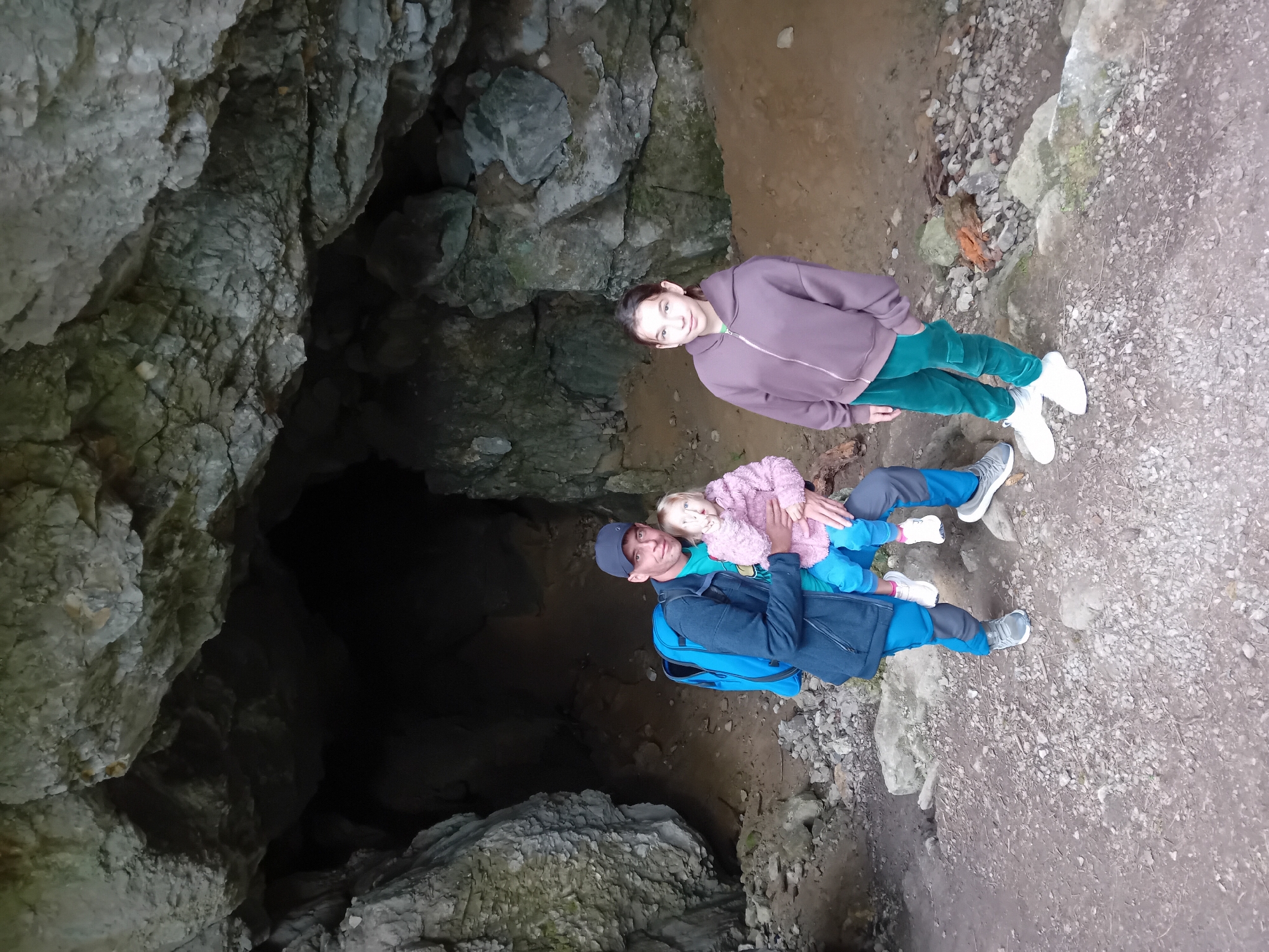 A family of three is posing for a photograph outside the entrance of a cave in Zuberec, Slovakia. The father, wearing a blue jacket and a baseball cap, is holding a young child in his arms. The child is wearing a pink knitted jacket. To the right of the father and child is a teenage girl wearing a purple hoodie and green pants. The cave entrance is dark and appears to be made of rock. The ground in front of the cave is covered in gravel and small rocks.