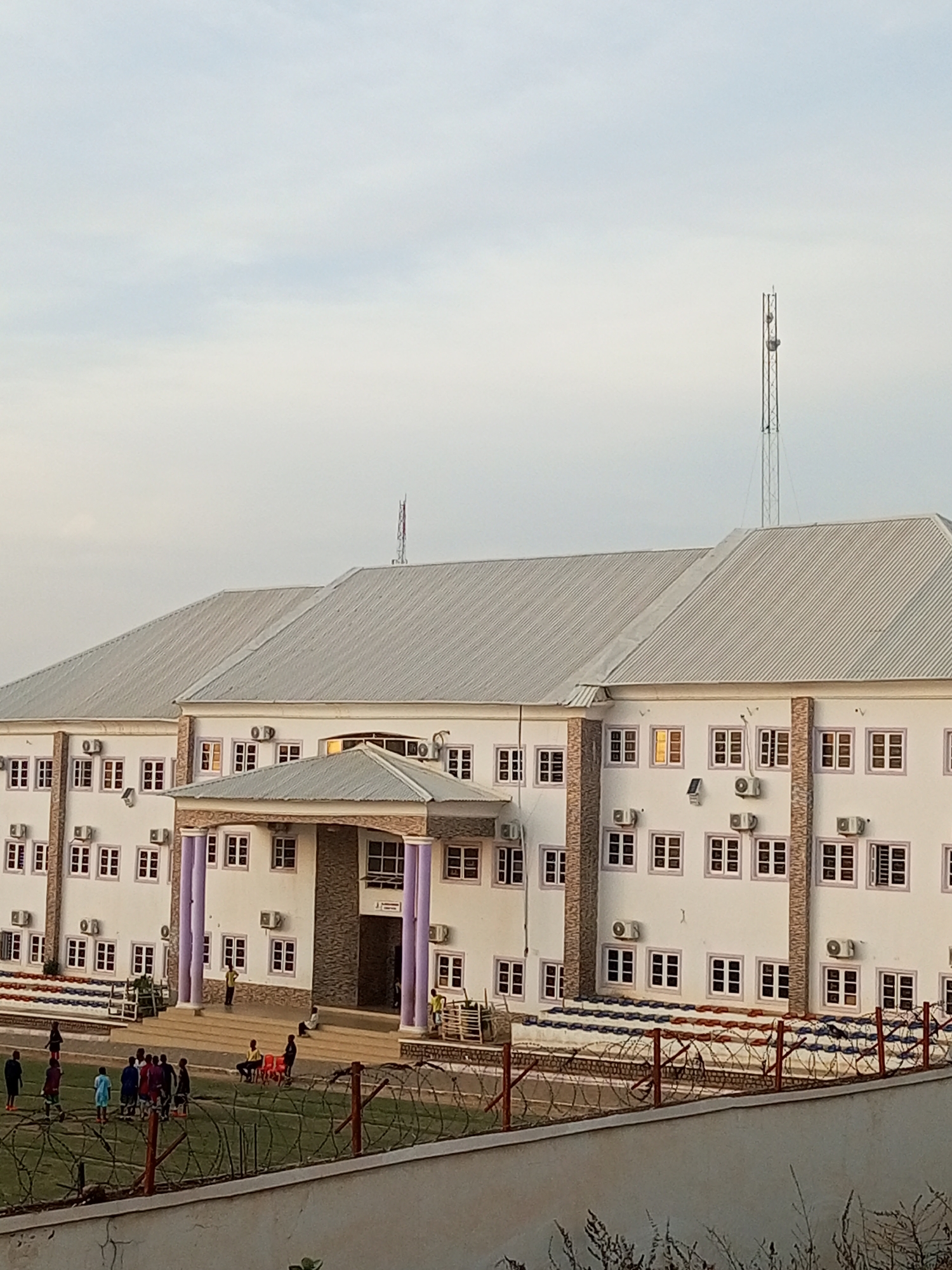 The image depicts a modern, multi-story building with a gray, corrugated metal roof, located in Gombe, Nigeria. The building's exterior is painted white, with a facade that includes numerous windows with white frames. Several air conditioning units are mounted on the exterior walls. The building features a prominent entrance with purple columns and a stone-clad portico. A telecommunications tower is visible in the upper right corner of the image, against a hazy sky.
In front of the building, a grassy field, likely a sports field, is visible. Several individuals, appearing to be young people, are present on the field, some engaged in activities that suggest a game or practice. There are also bleachers with red and blue seating in front of the building. A fence, reinforced with barbed wire, separates the field from the foreground. The overall environment suggests an institutional setting, possibly a school or a community center, within the city of Gombe.
