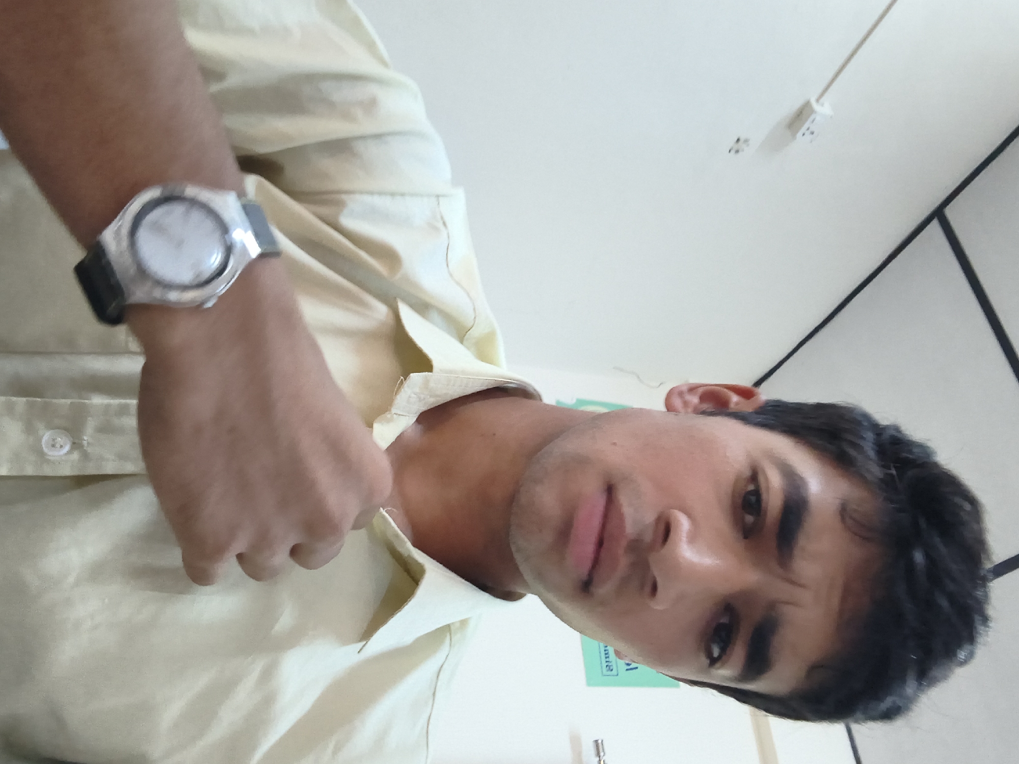 A young man with dark hair and a light-colored shirt is shown in a close-up, waist-up portrait. He is wearing a silver wristwatch on his left wrist. The background is a plain, light-colored wall with a white electrical outlet visible on the left side. The lighting suggests an indoor setting. There is no indication of the city of Dharamshala, India, in the image.
